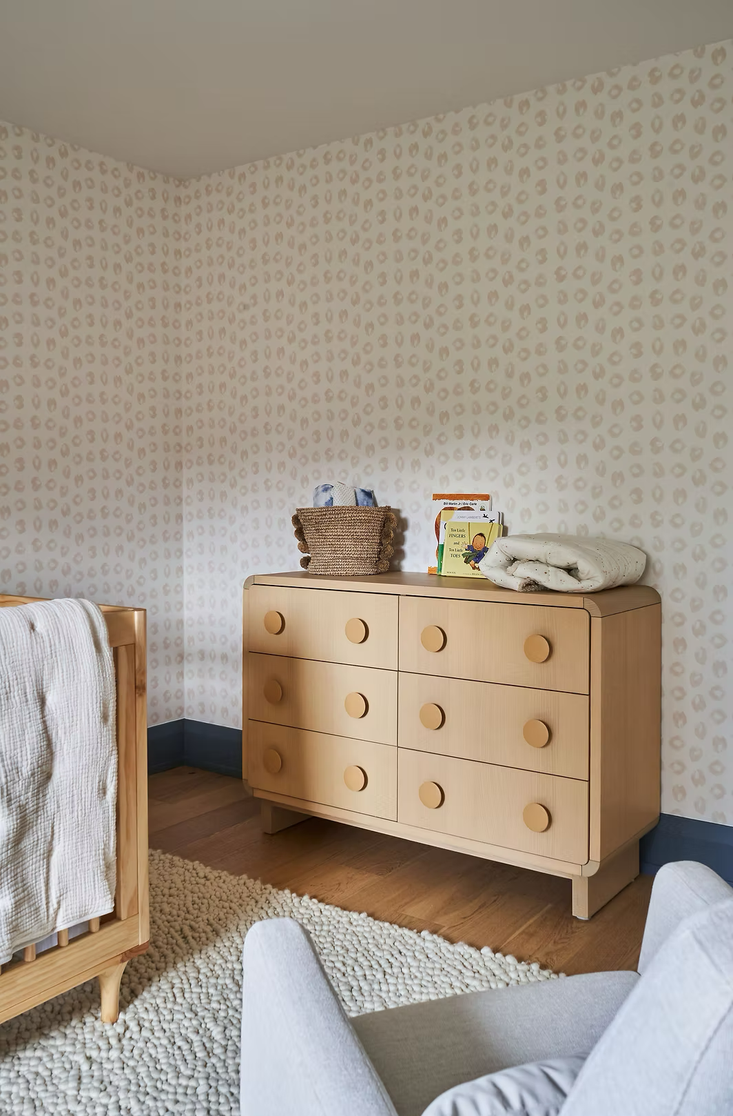 A light wood dresser with six drawers, topped with a woven basket, children's books, and a white blanket, in a bedroom with patterned wallpaper and a white textured rug.