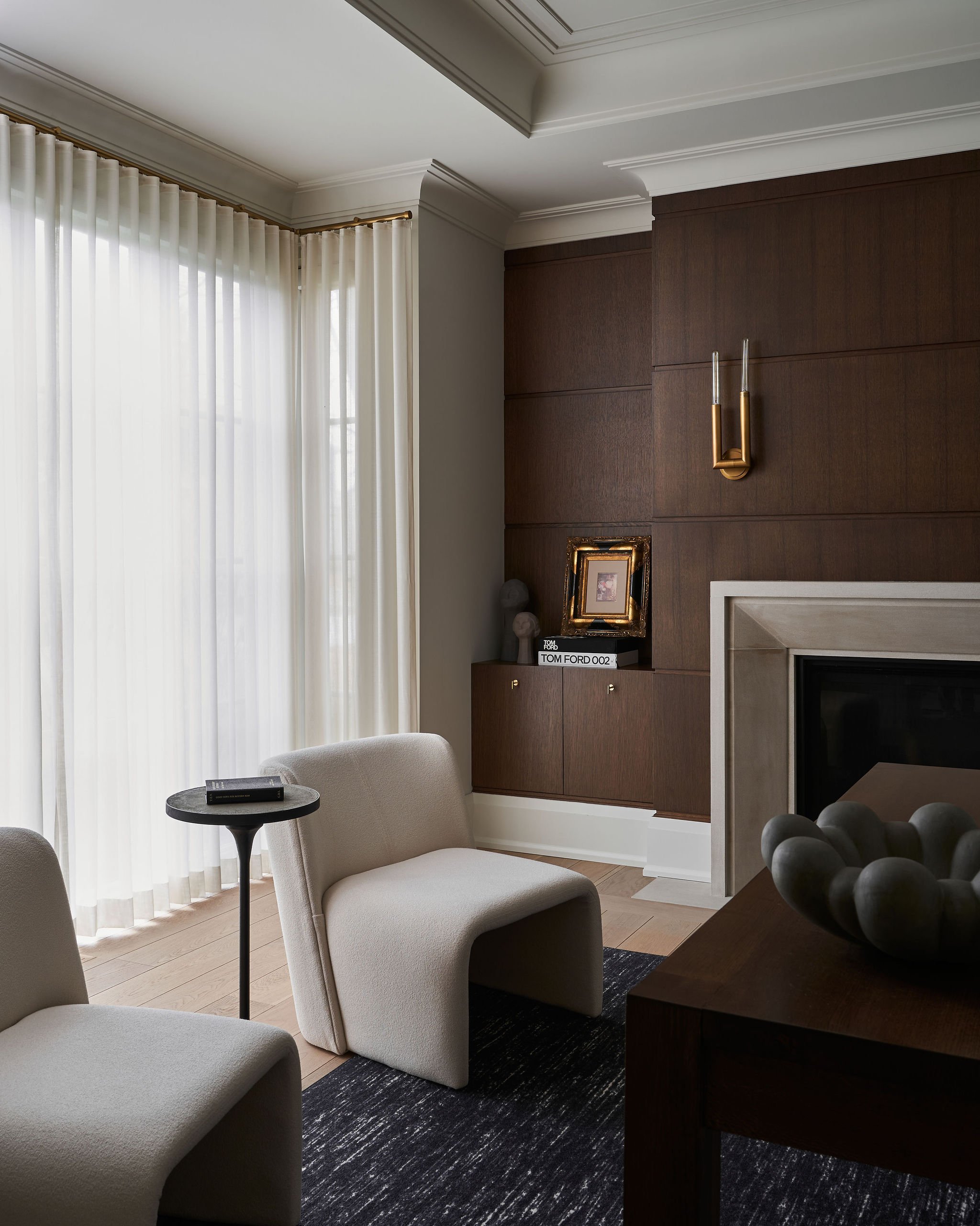 Living room with beige chairs, a small black side table, white curtains, a wooden wall with framed artwork, a modern gold and glass wall sconce, and a section of a fireplace with decorative dark spheres on a table.