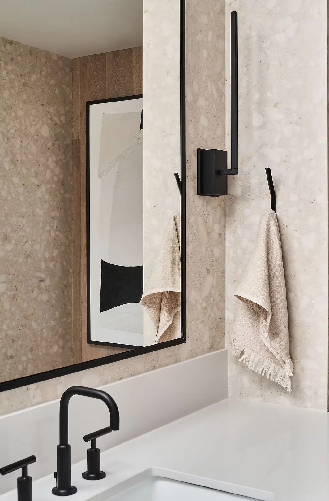 Bathroom mirror reflecting a beige towel hanging on a black hook, beige stone wall, and part of a modern black faucet over a white bathtub.