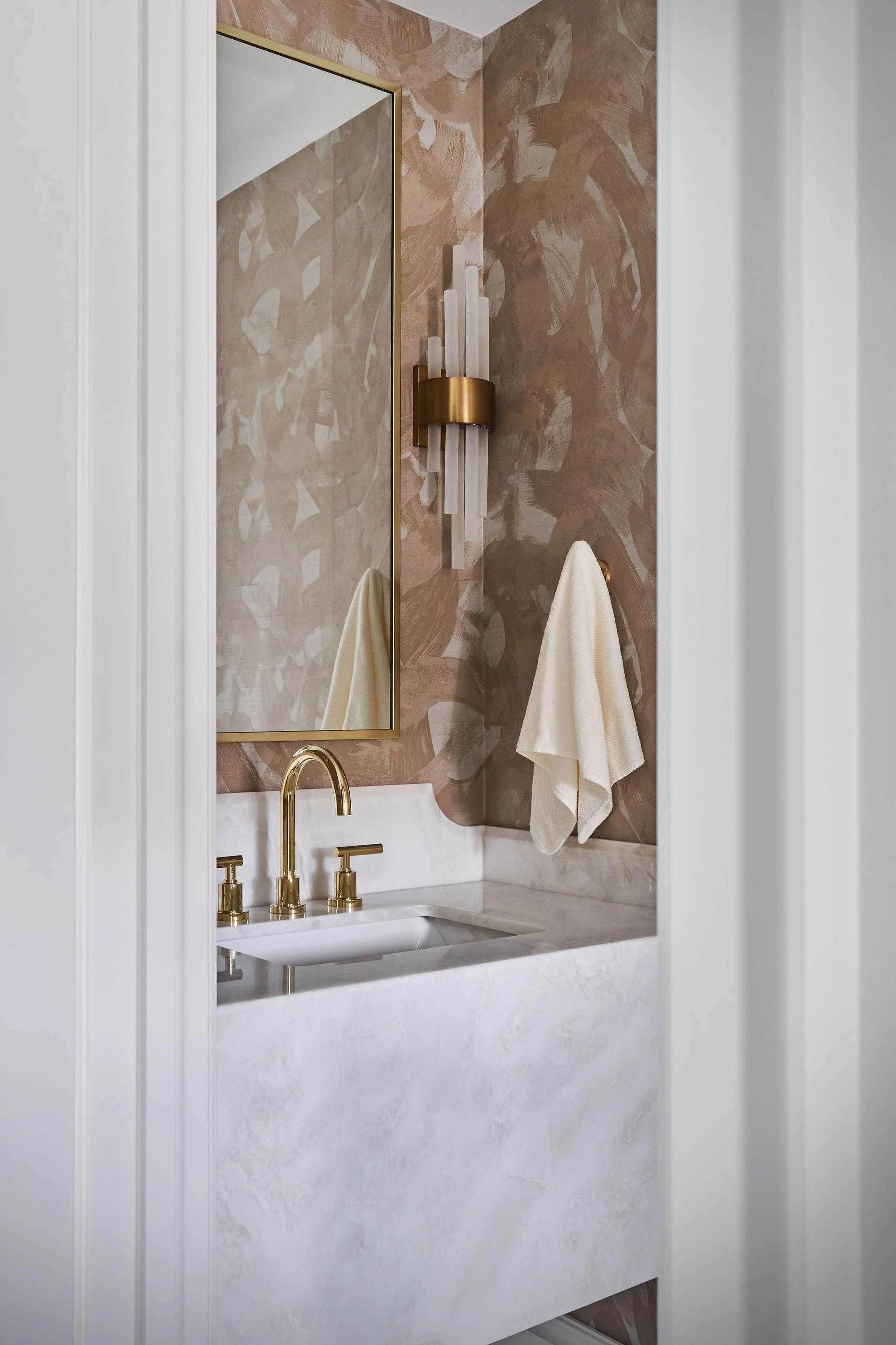 Bathroom sink with gold fixtures, a marble countertop, a large mirror, beige decorative wall panels, a wall-mounted sconce light fixture, and a towel hanging on a hook.