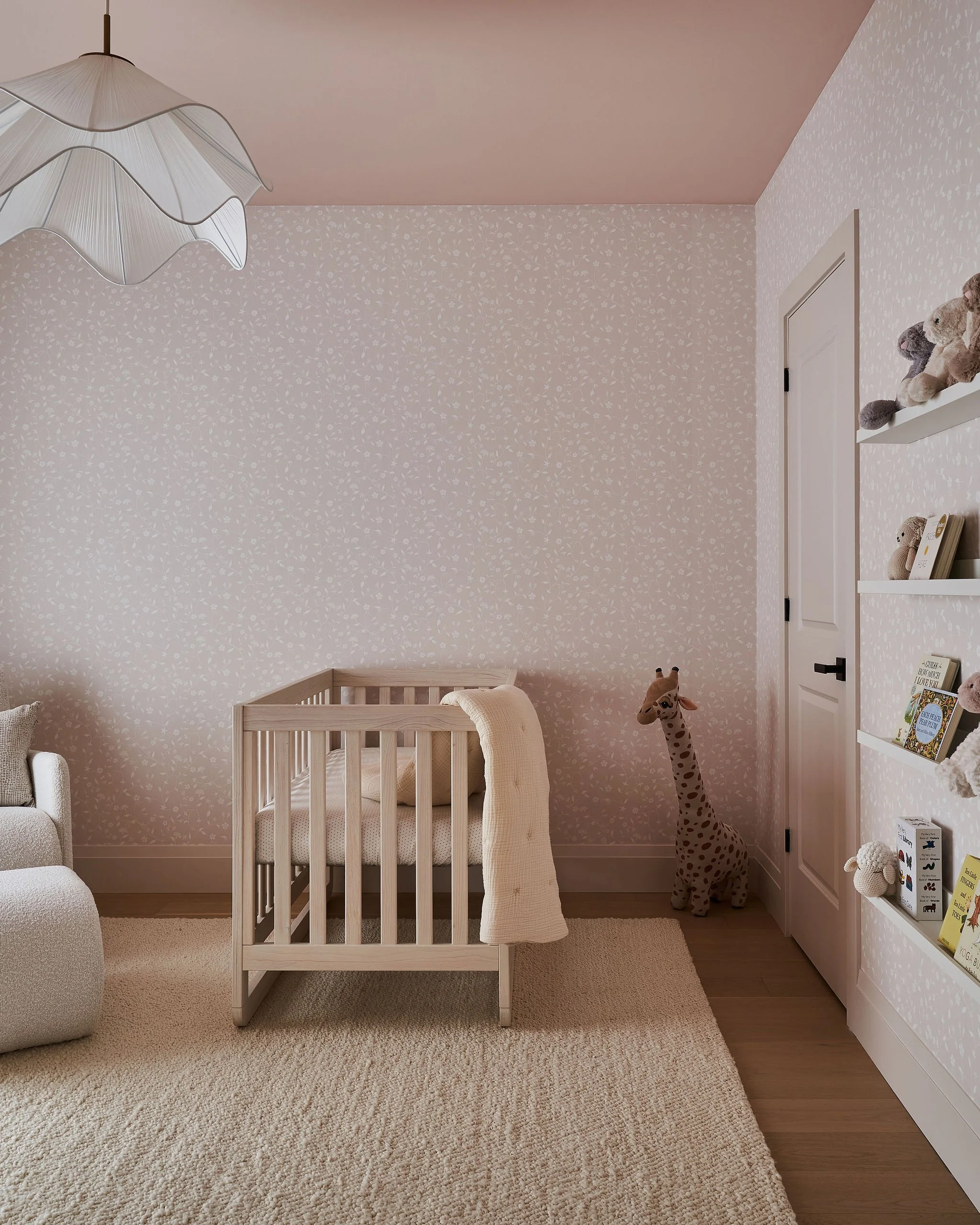 A nursery room with wallpapered walls, a white crib with a blanket hanging over the side, a stuffed giraffe toy, a white shelf with books and stuffed animals, and a textured beige rug on a wooden floor.