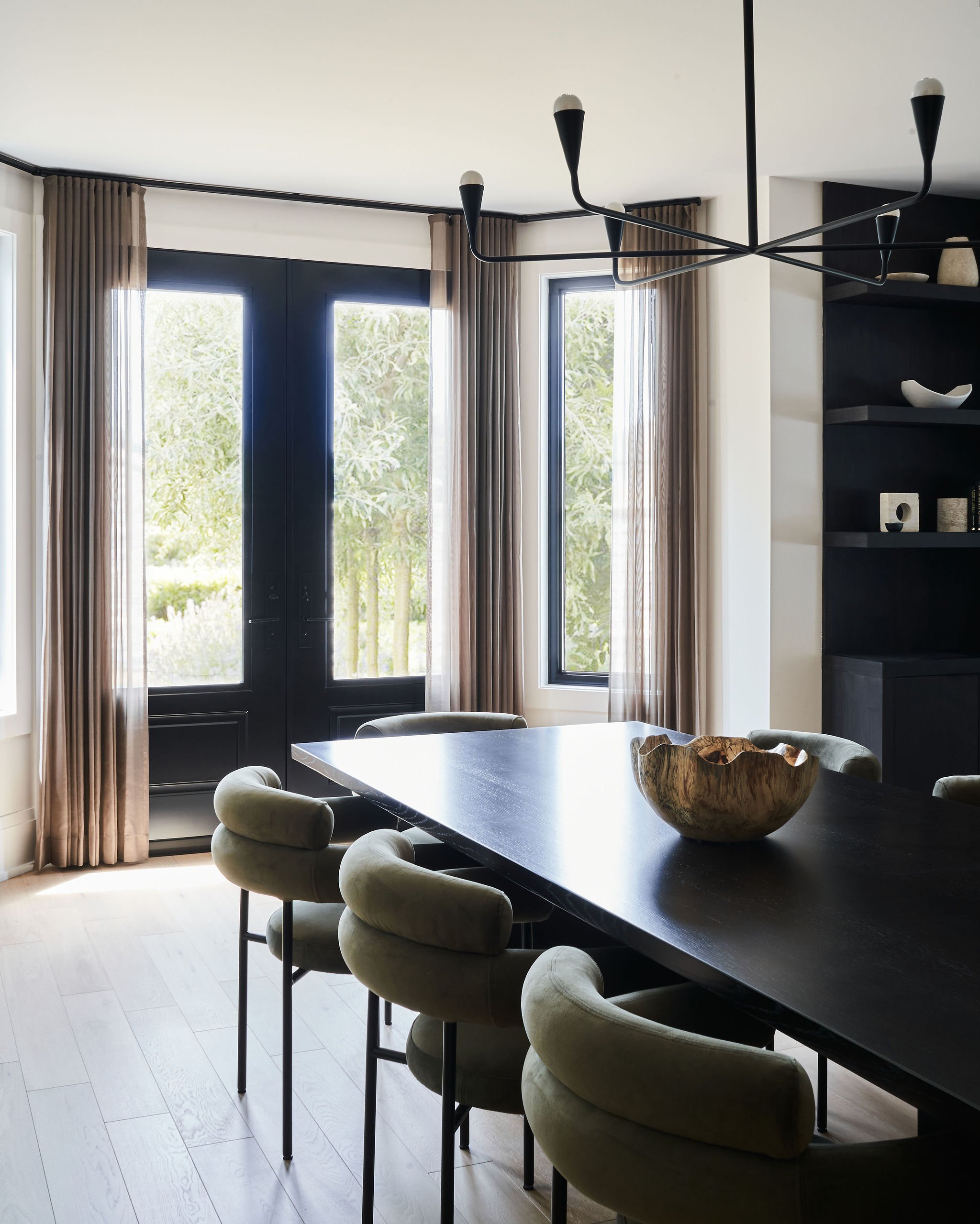 Modern dining area with window and black table, beige cushioned chairs, beige curtains, and a black chandelier with multiple arms
