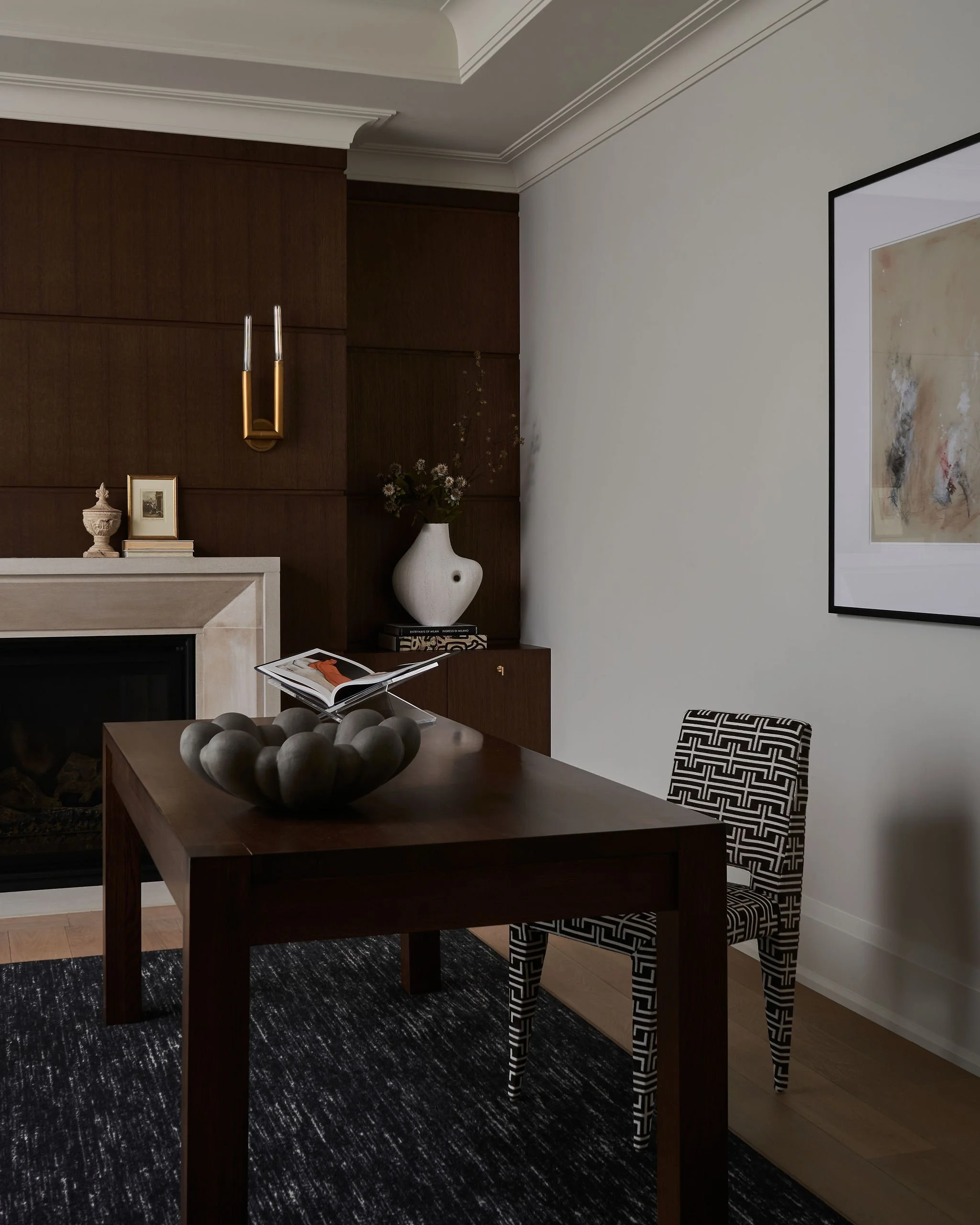 Living room with dark wood paneling, white ceiling, and white walls. A fireplace with a mantel holds decorative objects, including a framed photo, a small sculpture, and books. A large white vase with flowers is on a side table. A wooden table has a 