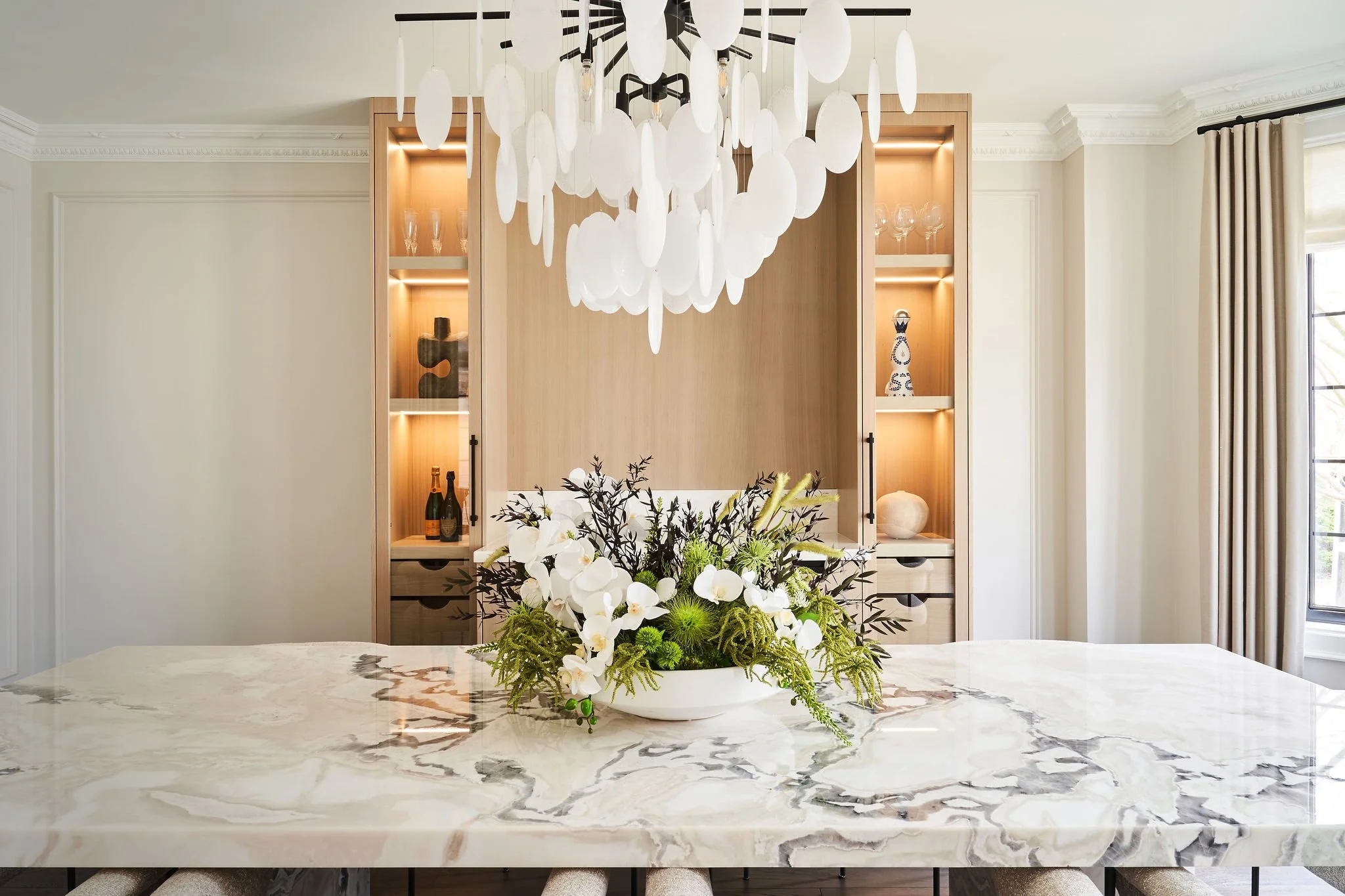 A modern kitchen or dining area with a marble countertop, floral centerpiece, wooden shelving with decorative items, and a white pendant light fixture hanging from a black ceiling mount.