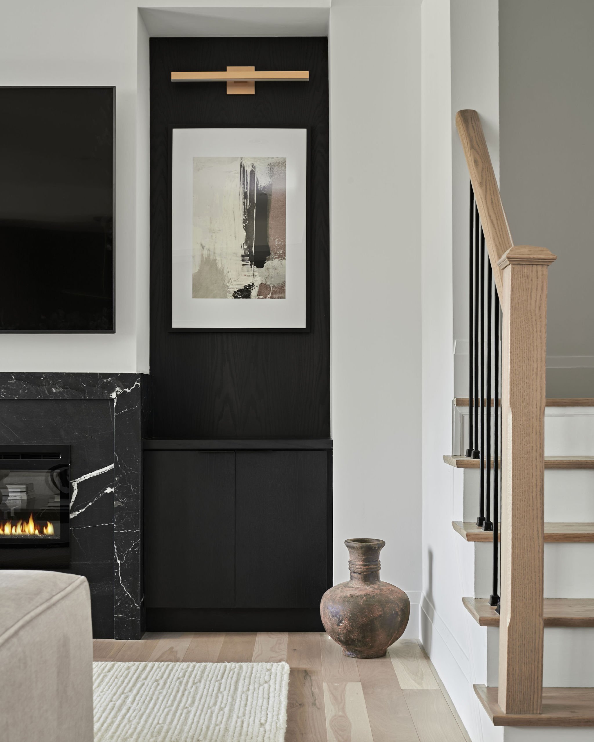 Interior view of a living space with a black and white fireplace, a beige armrest, a textured white rug, a large decorative ceramic pot, and a staircase with wooden handrail and black metal balusters.