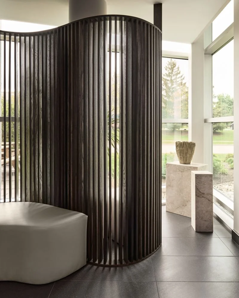 Interior space with a curved dark wooden slatted partition, a cream-colored curved bench, and a white marble pedestal with a beige decorative vase, near large floor-to-ceiling windows showing outdoor greenery.