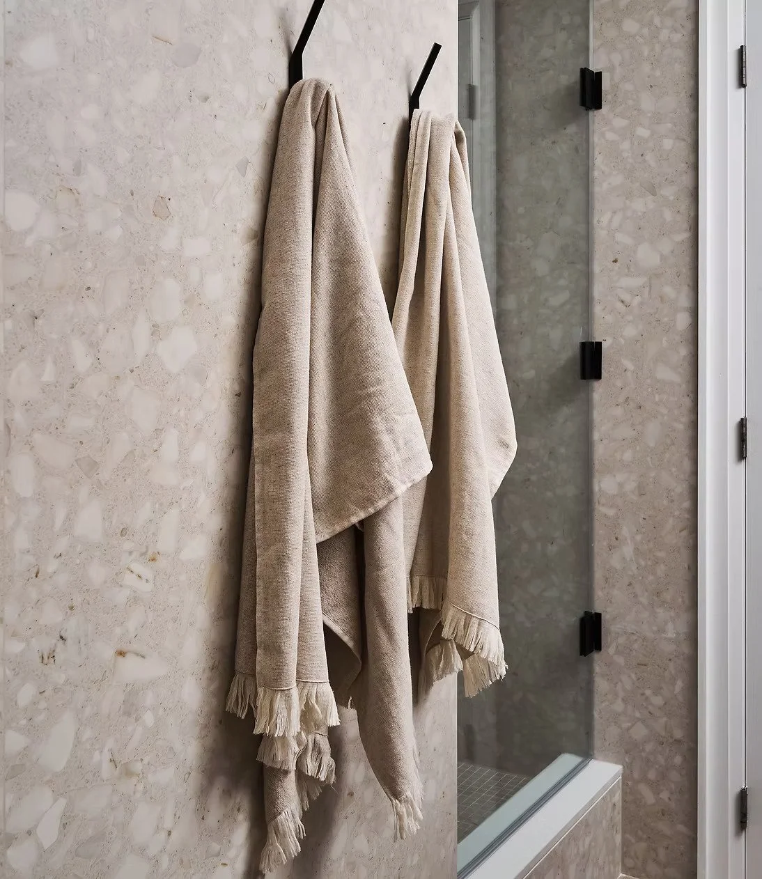 Two beige towels hanging on black hooks on a light textured wall in a bathroom, with a glass shower enclosure visible in the background.