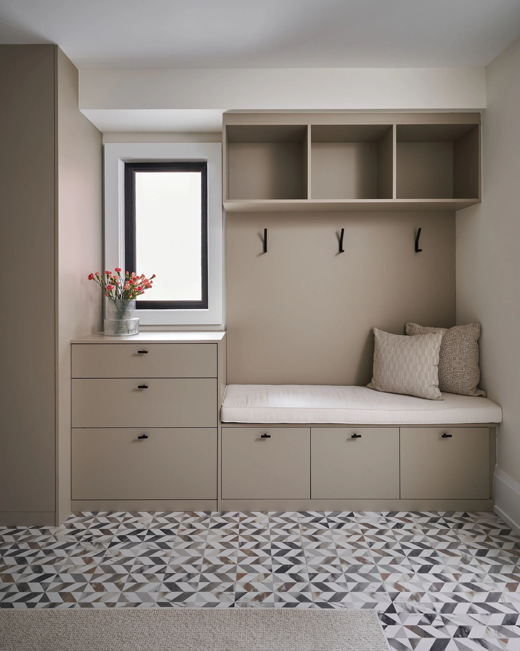 A minimalist entryway with a built-in storage bench featuring drawers, a wall-mounted shelf unit with three open cubbies, three black hooks beneath the shelf, a window with black trim, a vase with pink flowers on the window ledge, and a patterned til