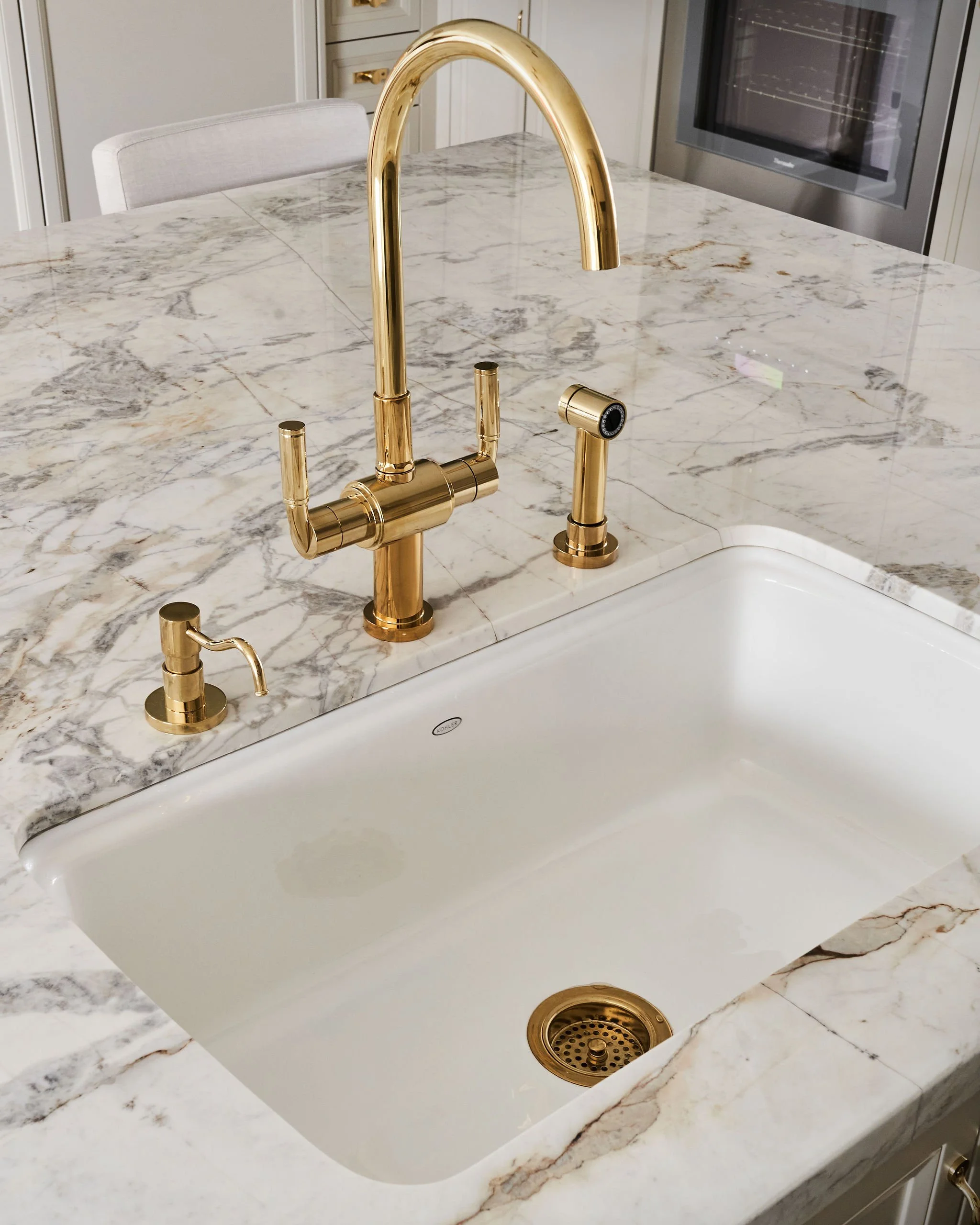A kitchen with a white marble countertop and a gold faucet. A white sink is visible underneath the countertop and a stainless steel oven is in the background.