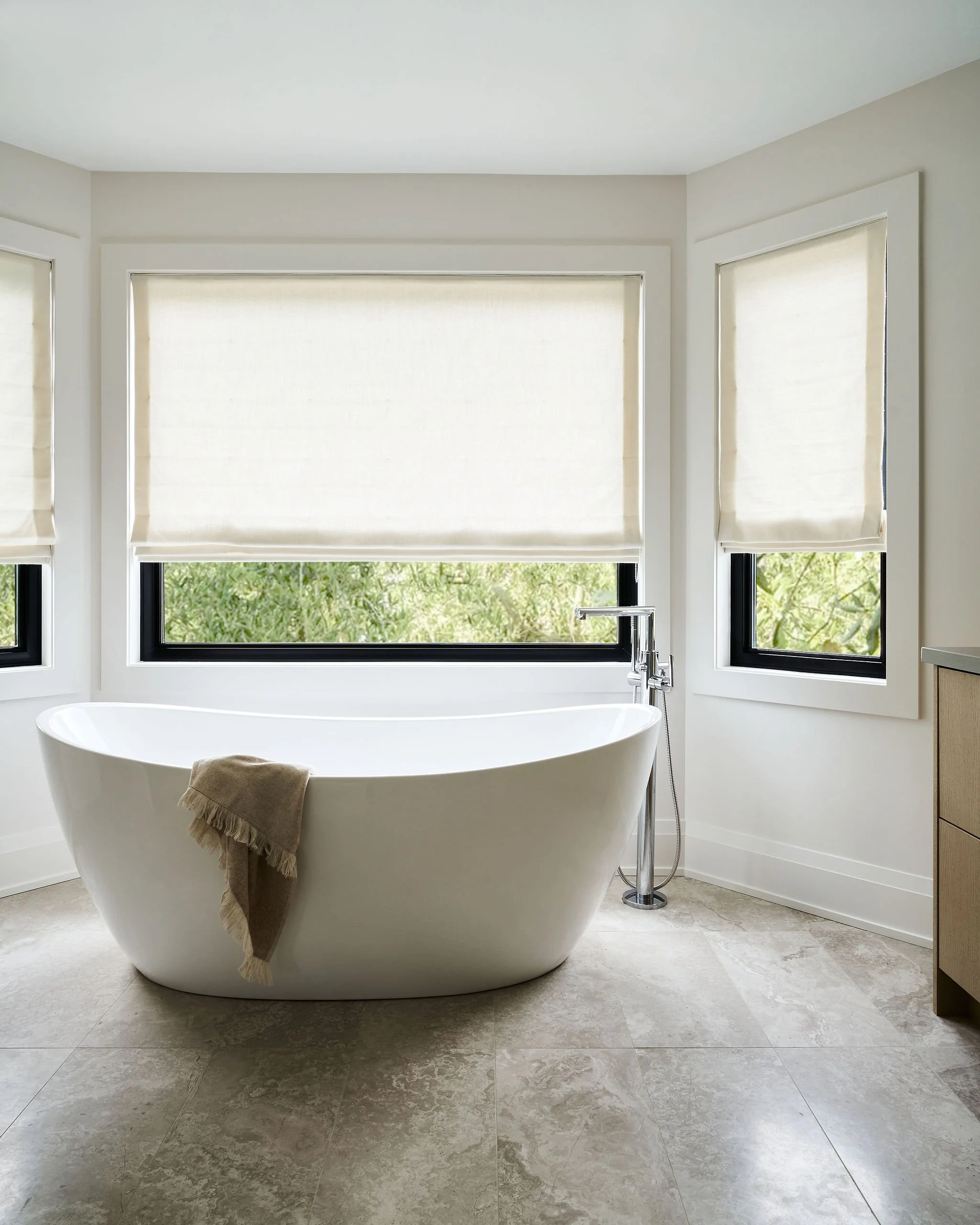Contemporary bathroom with a free-standing white bathtub, a beige towel draped over the edge, large windows with white blinds, beige tile flooring, and natural light.