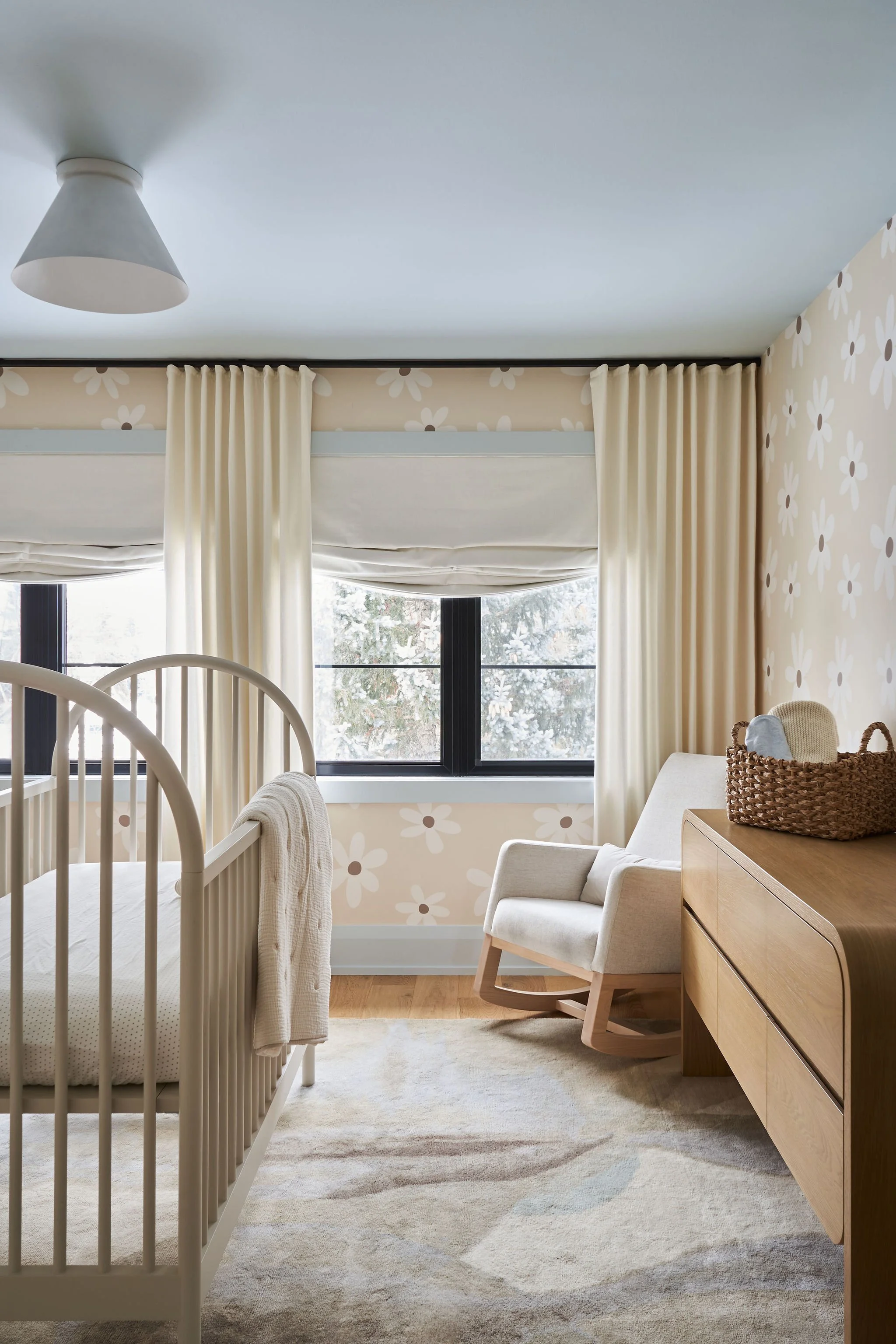 A cozy room with a white crib, beige curtains, a beige armchair, a wooden dresser, and wallpaper with white flowers on a beige background, illuminated by natural light from large windows.