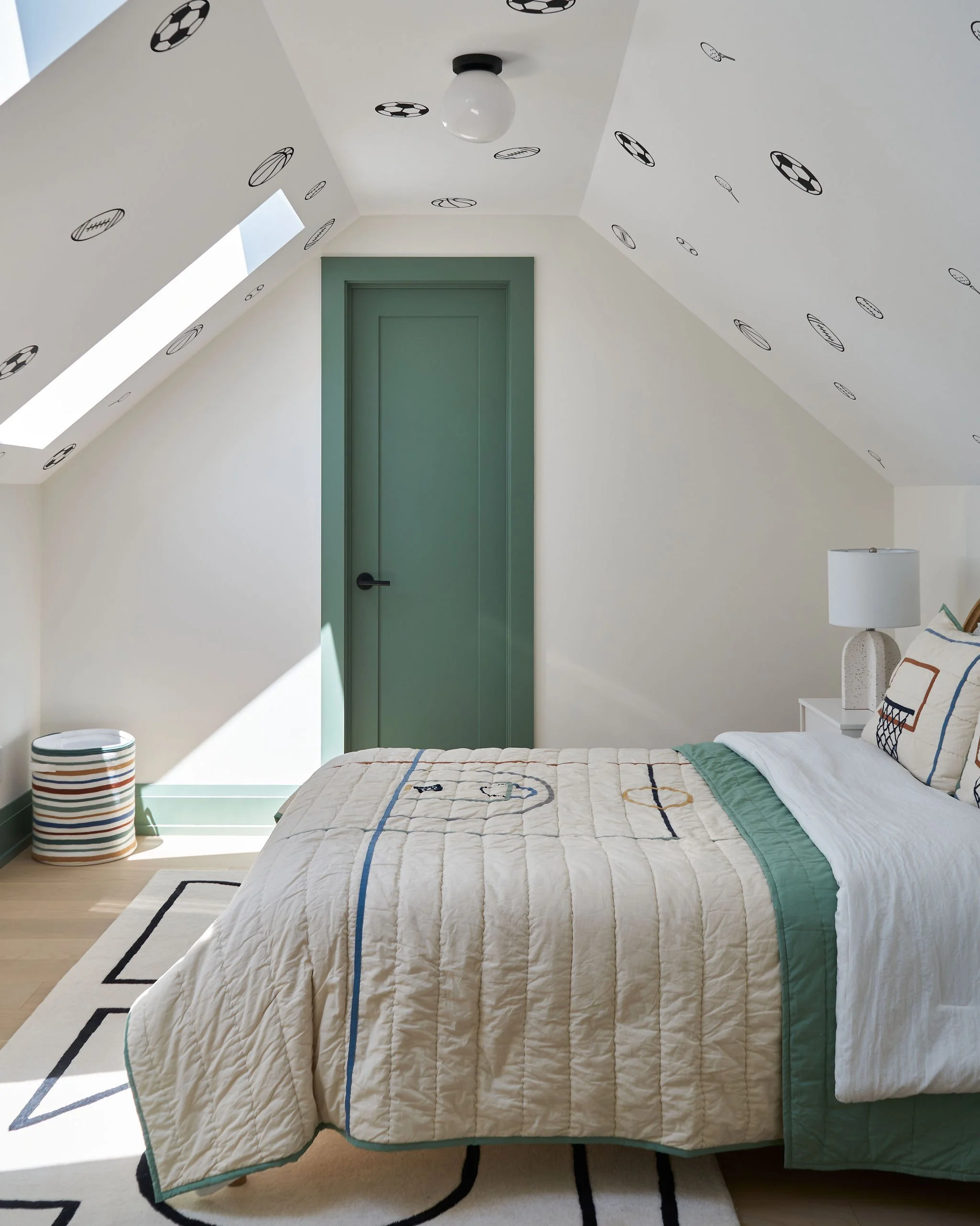 A bedroom with a bed, a green door, a patterned rug, a striped laundry basket, a table lamp, and a quilted bedspread with a tennis court design, in front of angled walls with tennis ball decals, all illuminated by natural light from a skylight.