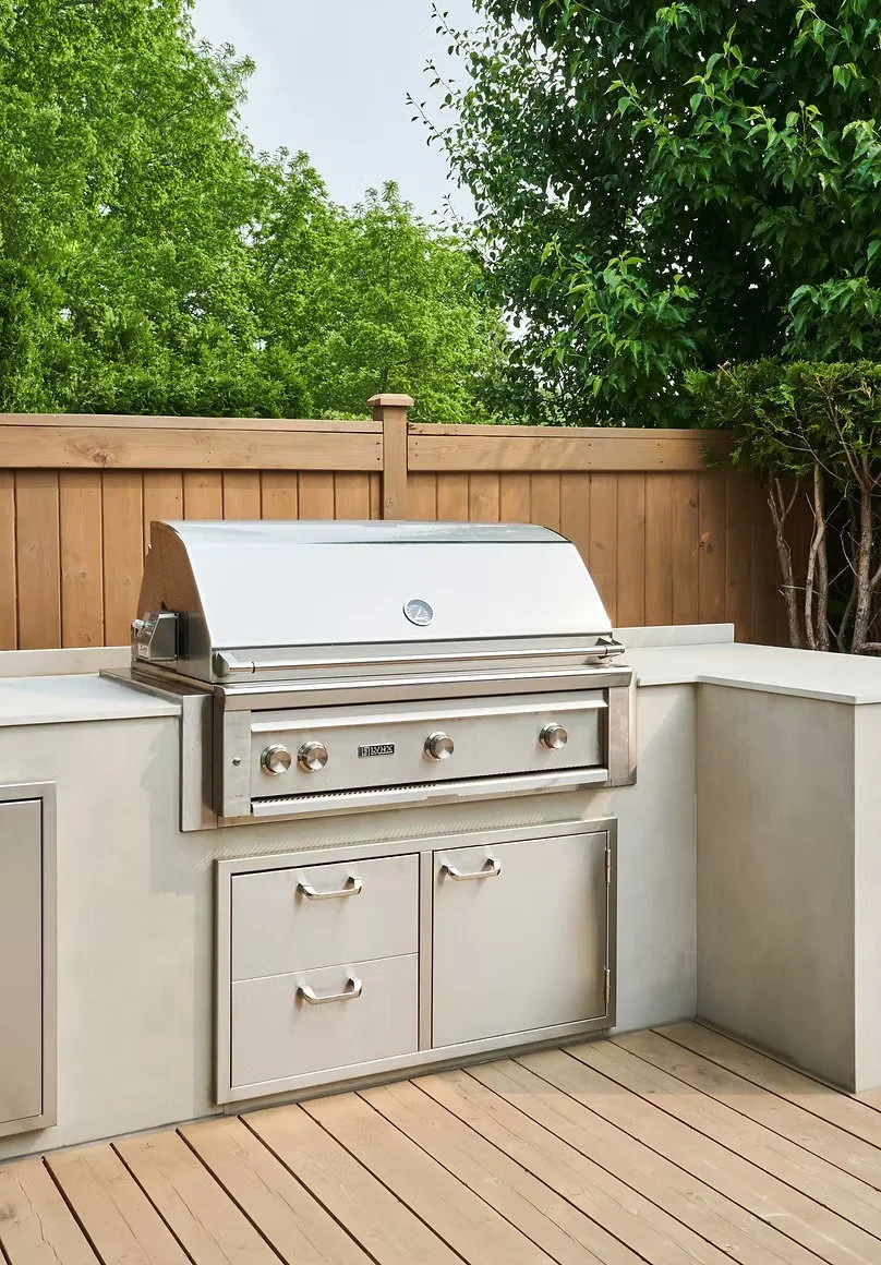 Outdoor kitchen with a stainless steel grill built into a white countertop, wooden fence with green trees in the background, and a wooden deck floor.