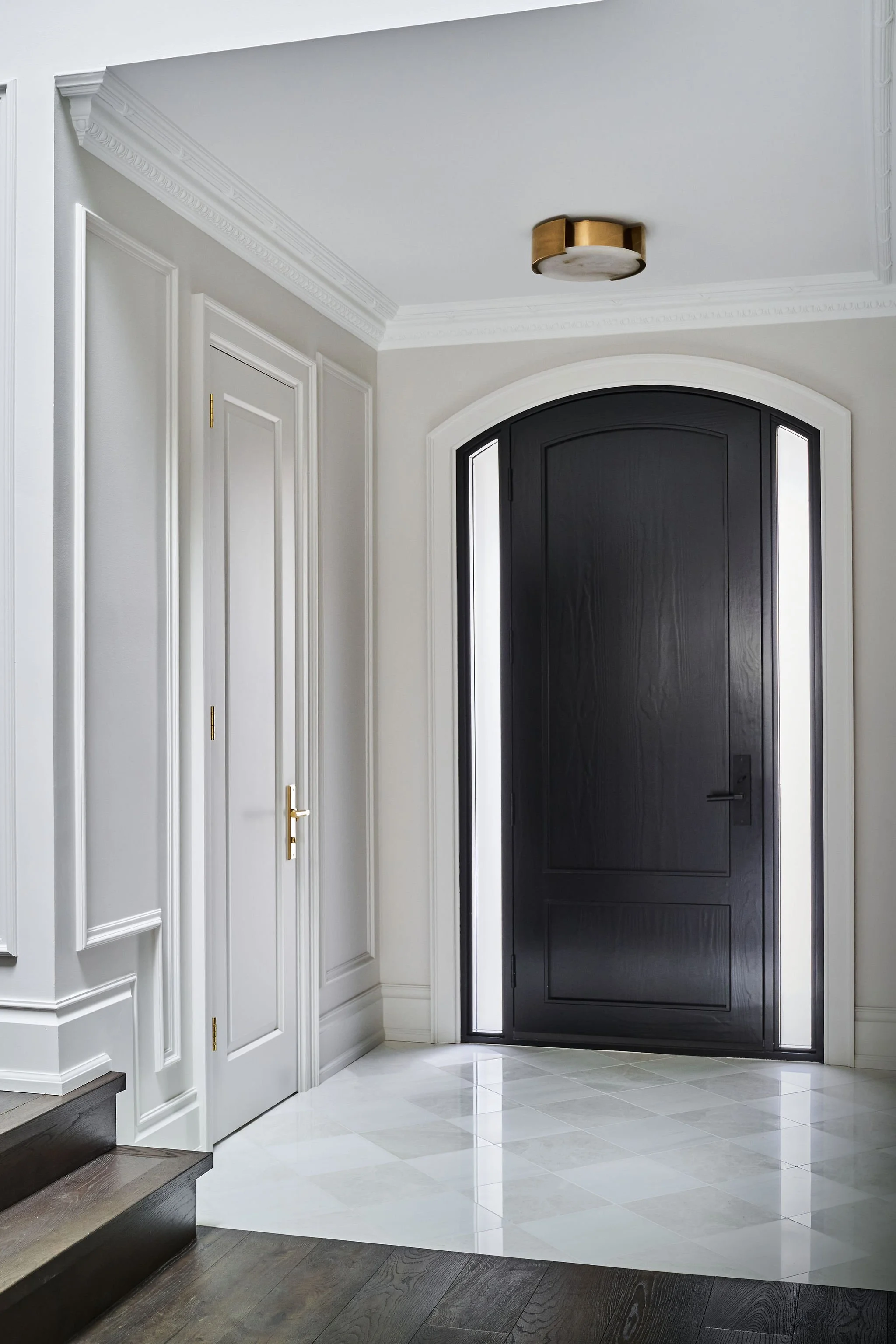 Inside a modern house entryway featuring a black arched front door with side windows, white crown molding, white wainscoting, white walls, and a gold/white ceiling light fixture. The floor has white and light gray tiles, and there are dark wooden ste