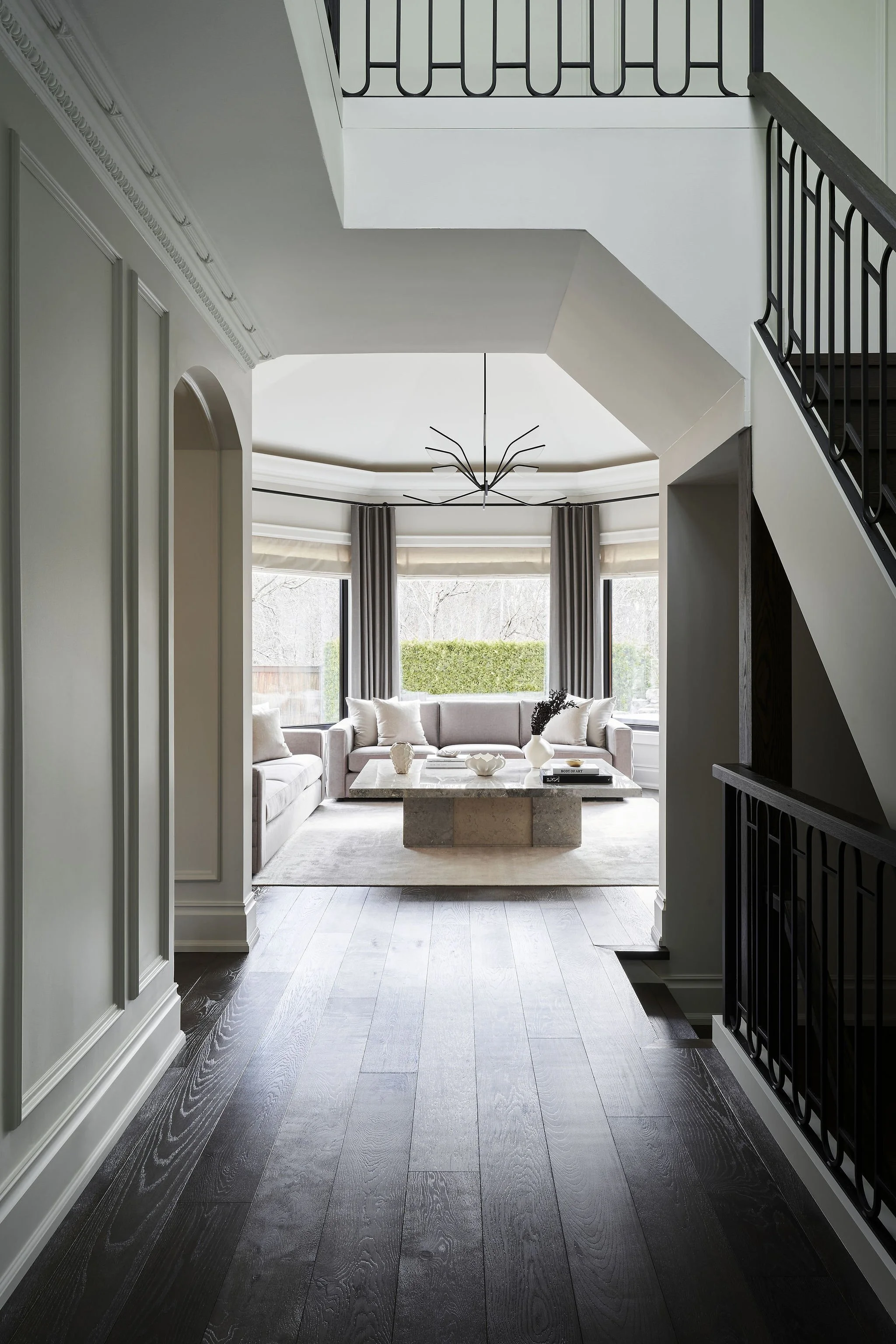 View of a living room with a light-colored sofa, coffee table, large windows with curtains, and modern lighting fixture. The room is bright with natural light, and the walls are painted a light color.
