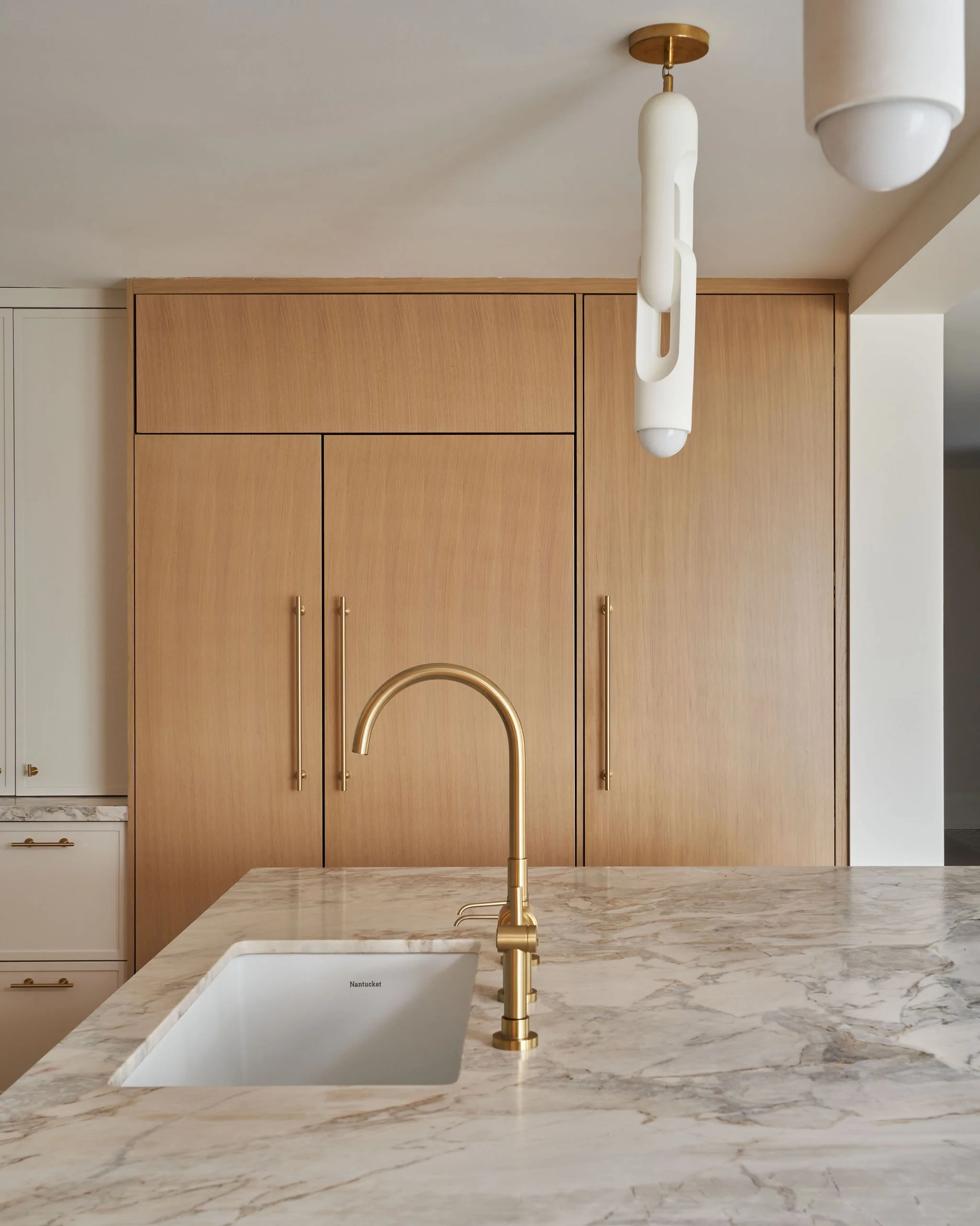 A modern kitchen with a marble countertop, a built-in white sink with the word 'Nantucket' inside, and a gold faucet. There are wooden cabinets with gold handles and a unique white and gold hanging light fixture above. The background includes a light