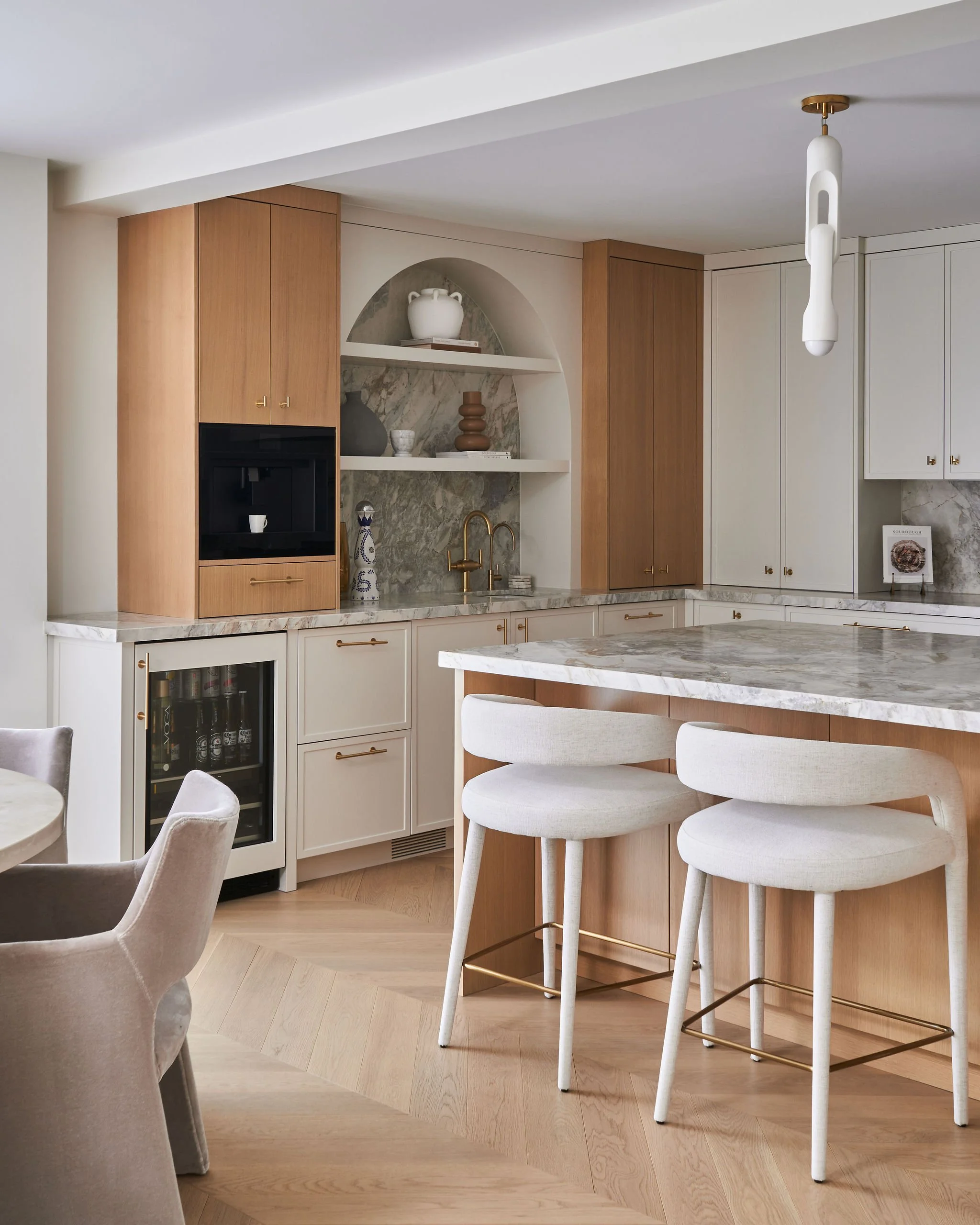 Modern kitchen with white and wood cabinets, marble countertops, gold fixtures, and a bar seating area with white stools.