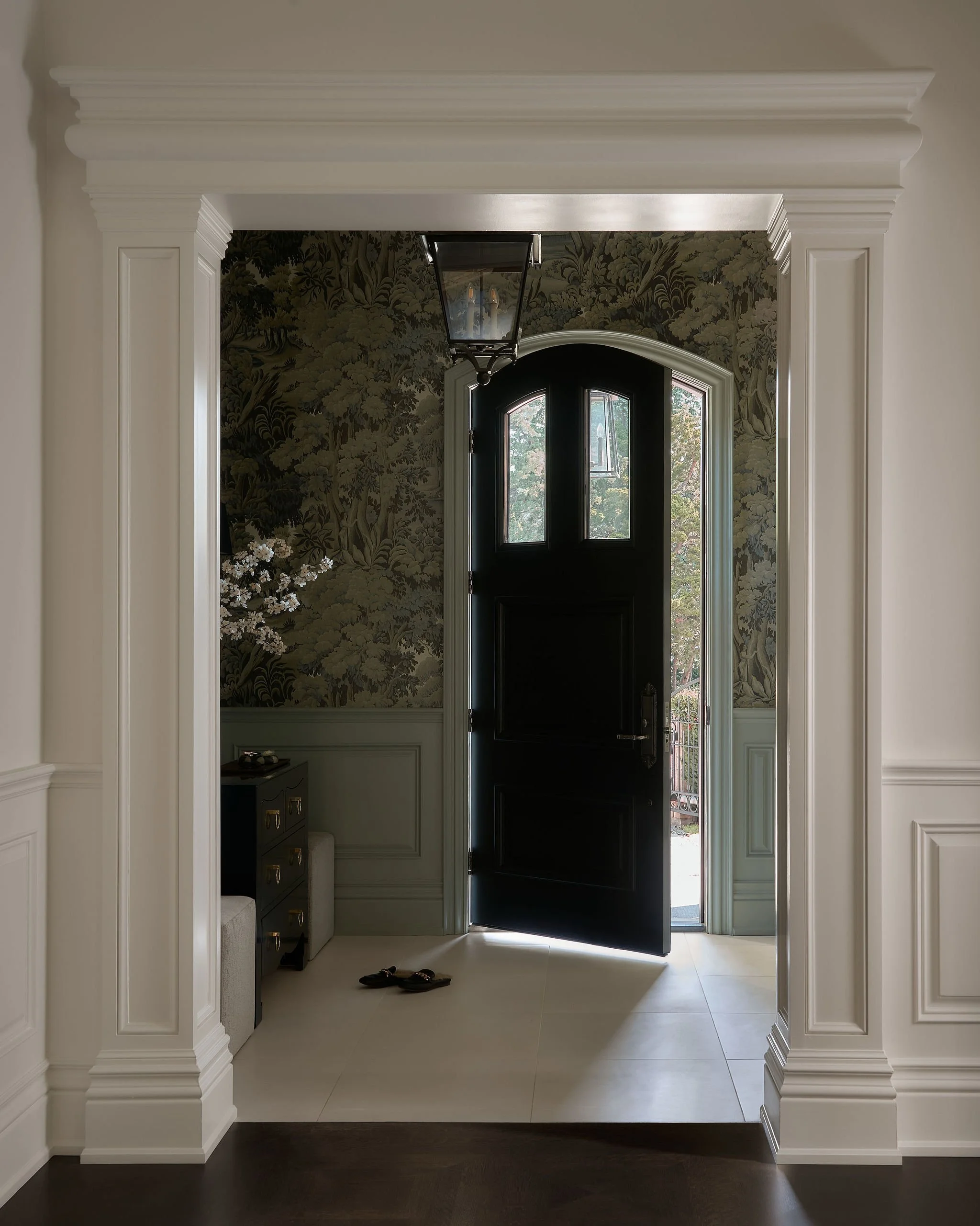 View of a front entrance door with black paint, framed by ornate white molding and trim, with a window and greenery visible outside. Inside, a pair of sandals rest on the floor near a small cabinet and floral wallpaper on the wall.