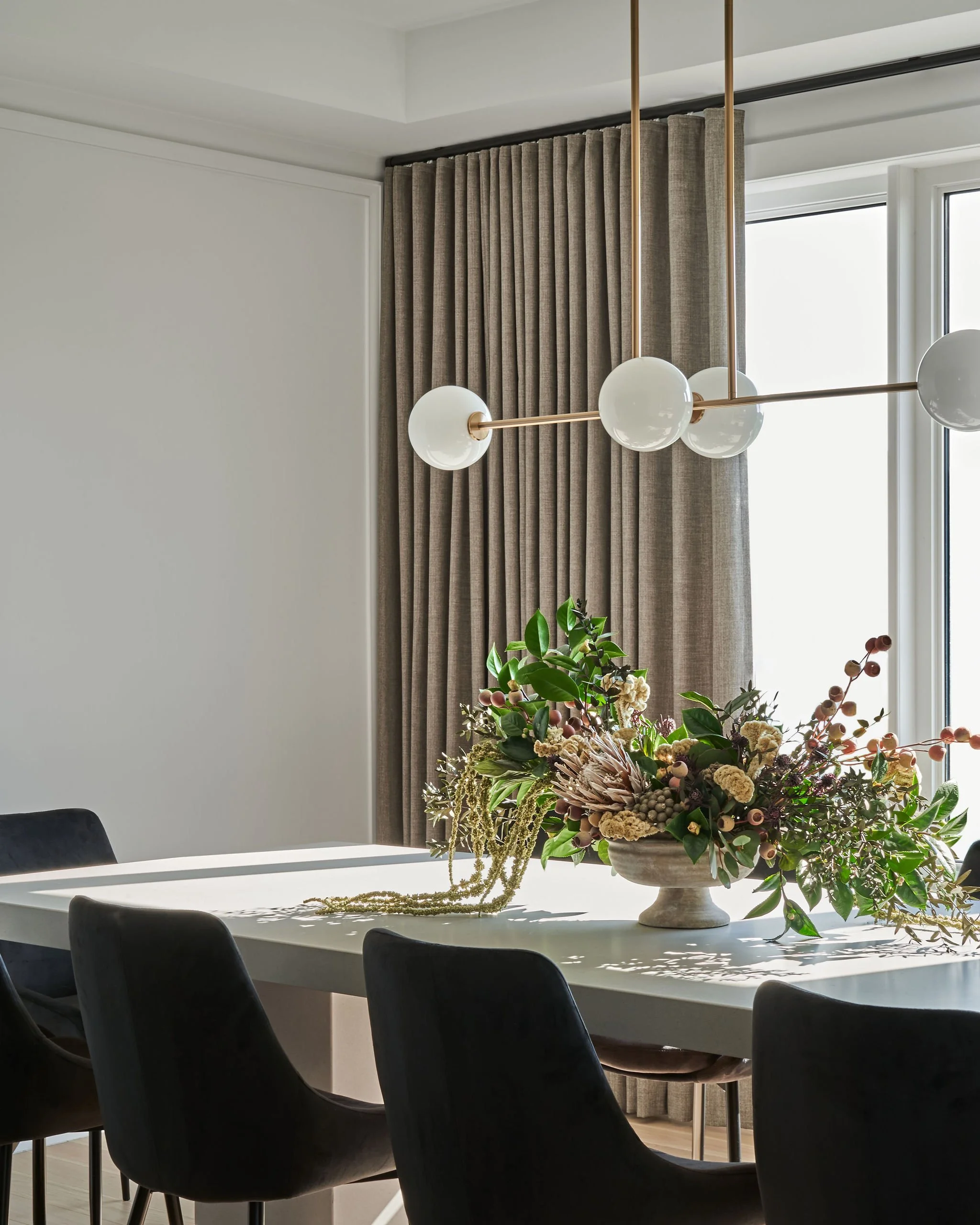 Modern dining room with a white table, black chairs, a floral arrangement in a large white vase, beige curtains, and a window with natural light.