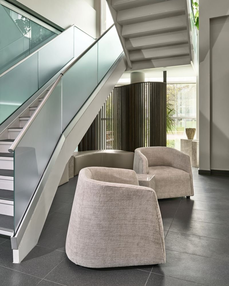Modern interior space with beige upholstered chairs, a curved window area, and a staircase with glass railing.