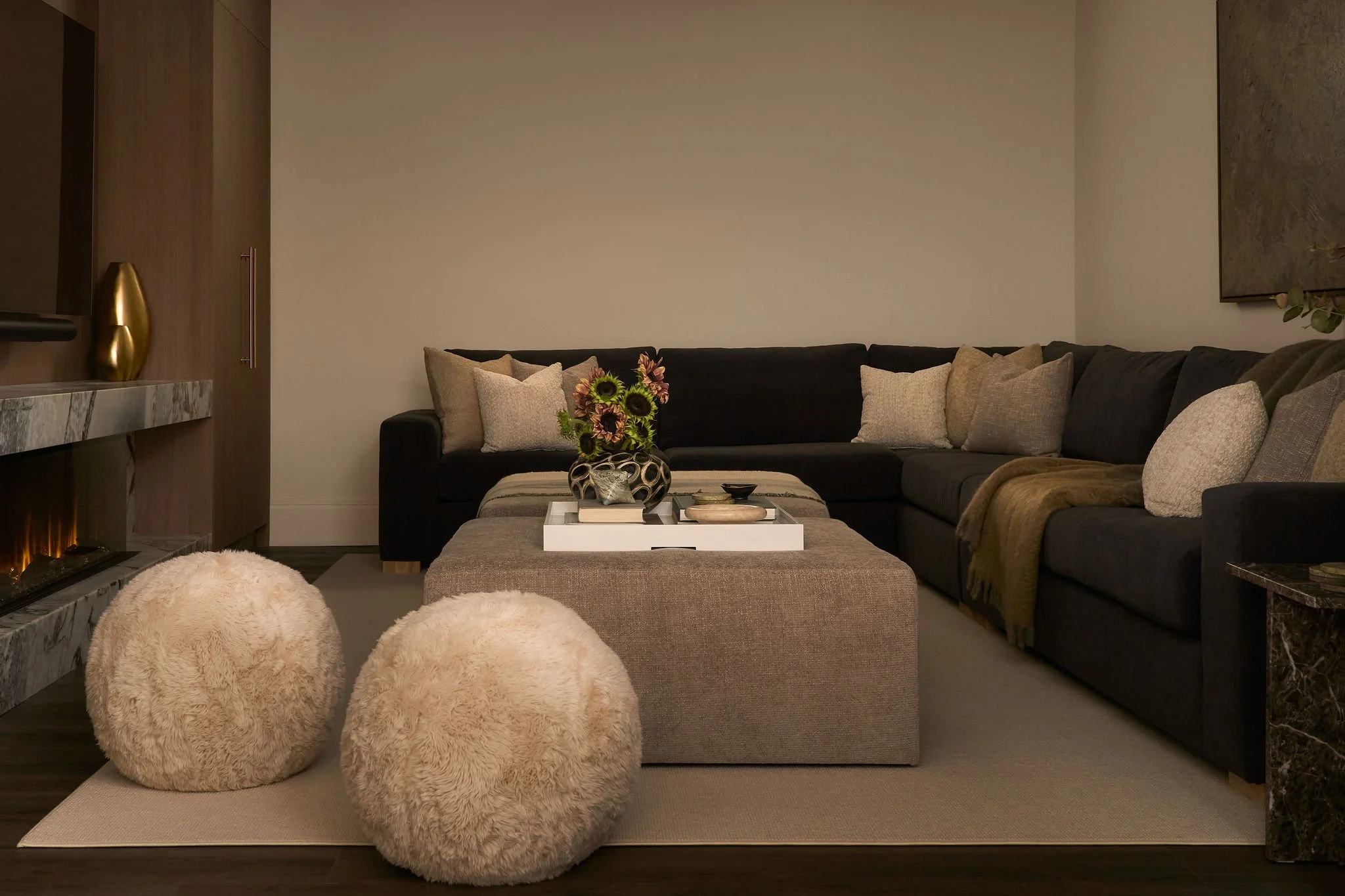 Living room with a dark gray sectional sofa, numerous beige and light brown pillows, a cream ottoman with a flower arrangement, a marble fireplace, and wall decorations.