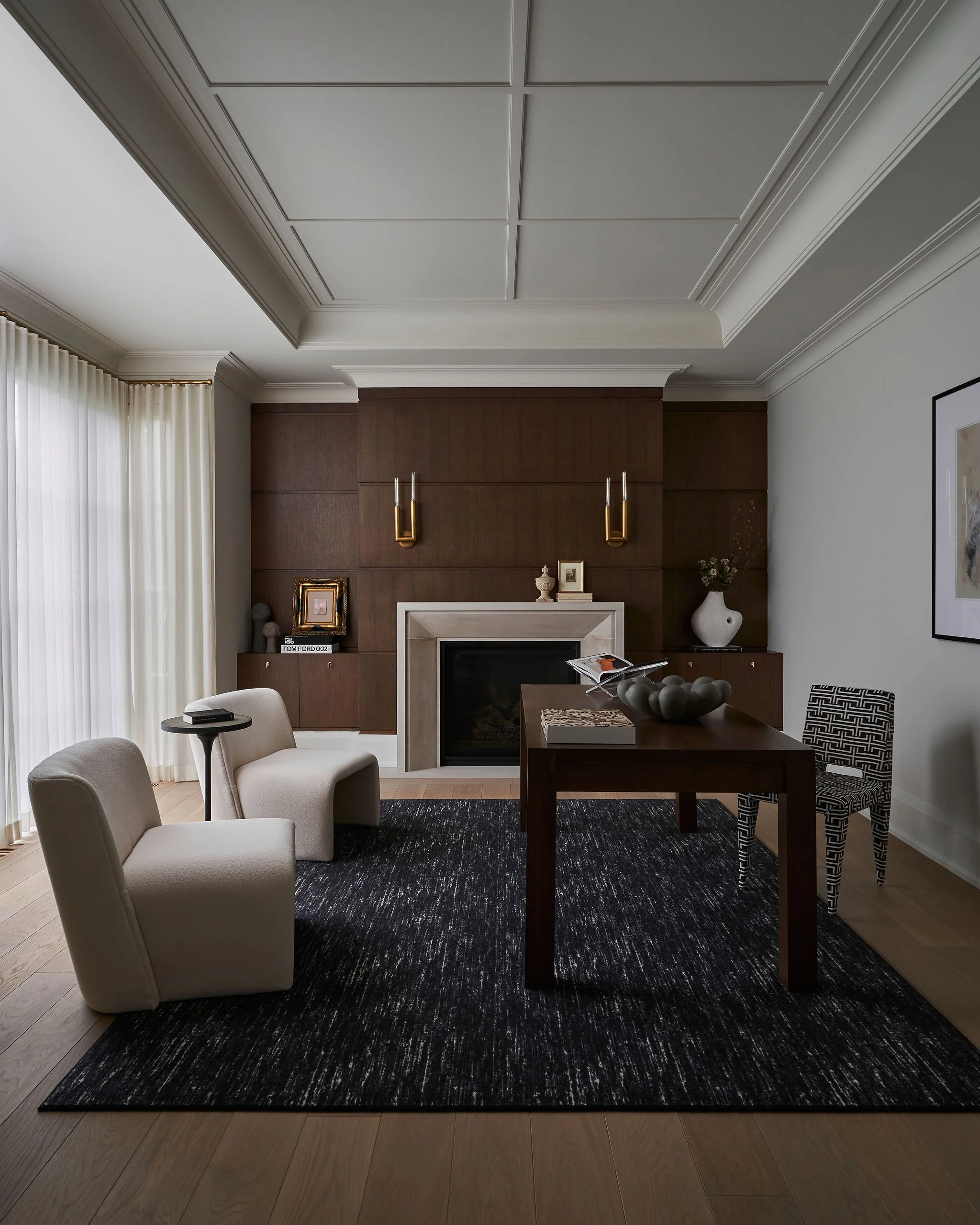 Interior view of a living room with a dark wood fireplace mantel, two wall-mounted gold light fixtures, a wooden dining table with decorative centerpiece and books, beige armchairs, black and white patterned chair, black area rug, and white curtains 