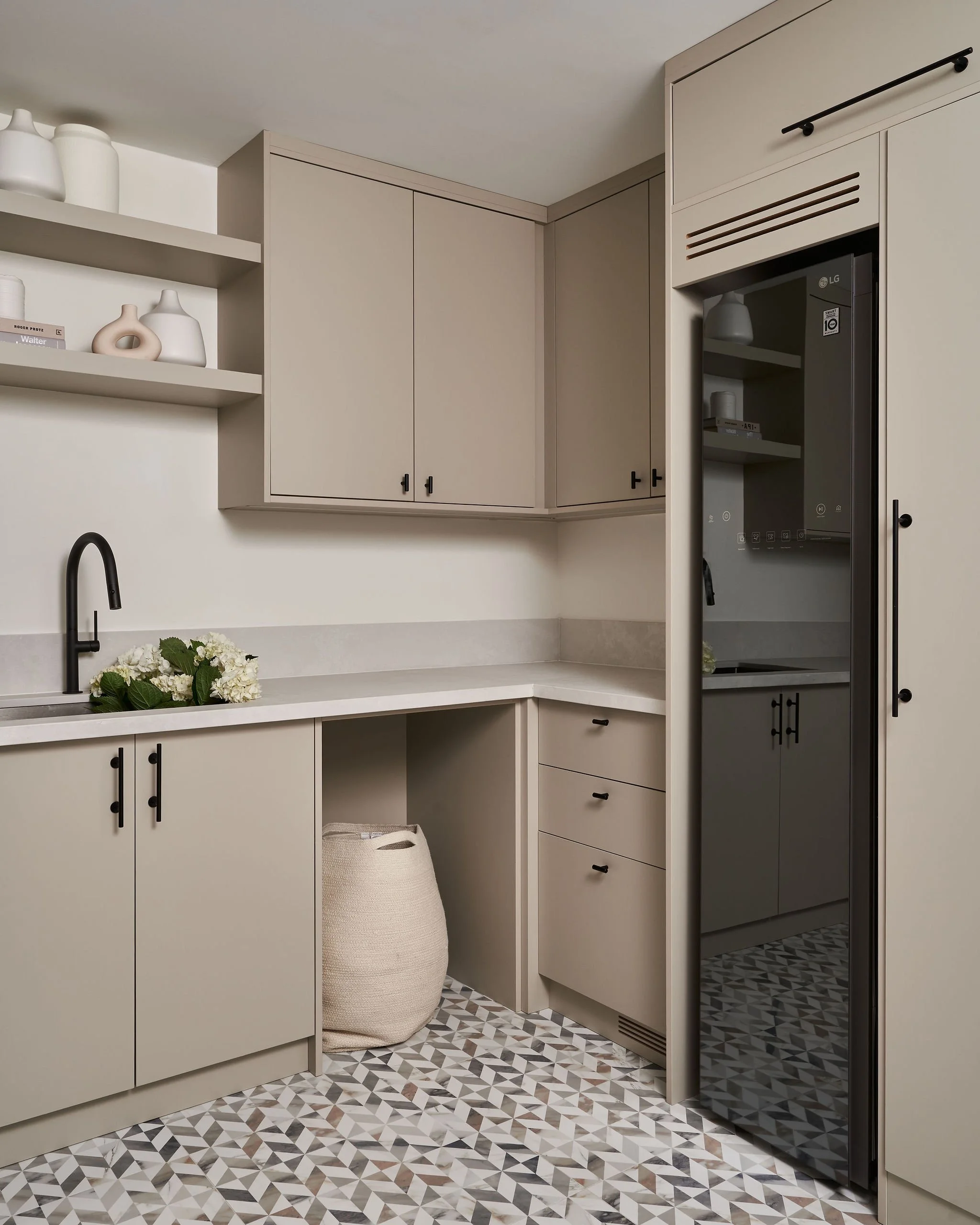 Modern kitchen with beige cabinets, black handles, patterned tile floor, black sink faucet, and open shelving with decorative vases and books, along with a large woven basket.