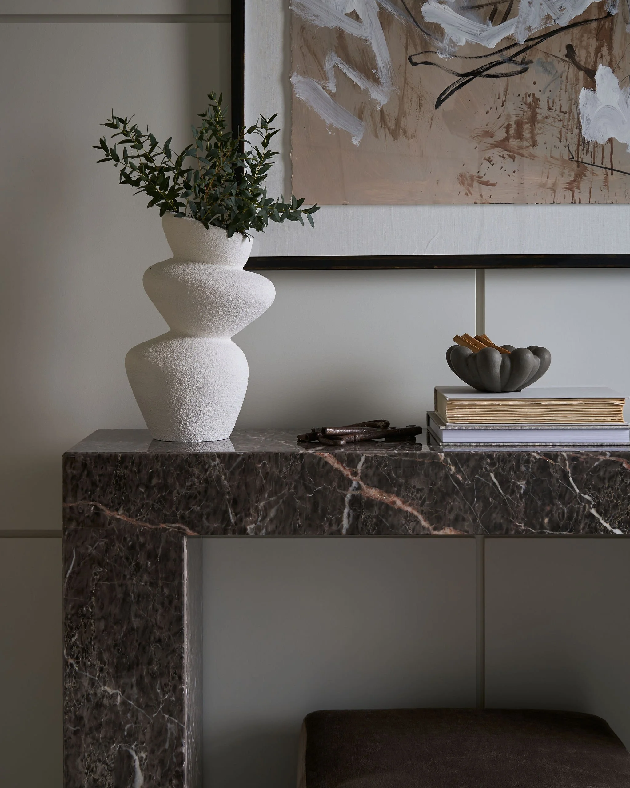 A decorative interior space featuring a white textured vase with green foliage, abstract artwork on the wall, dark marble table with a small sculpture, and a stack of books.
