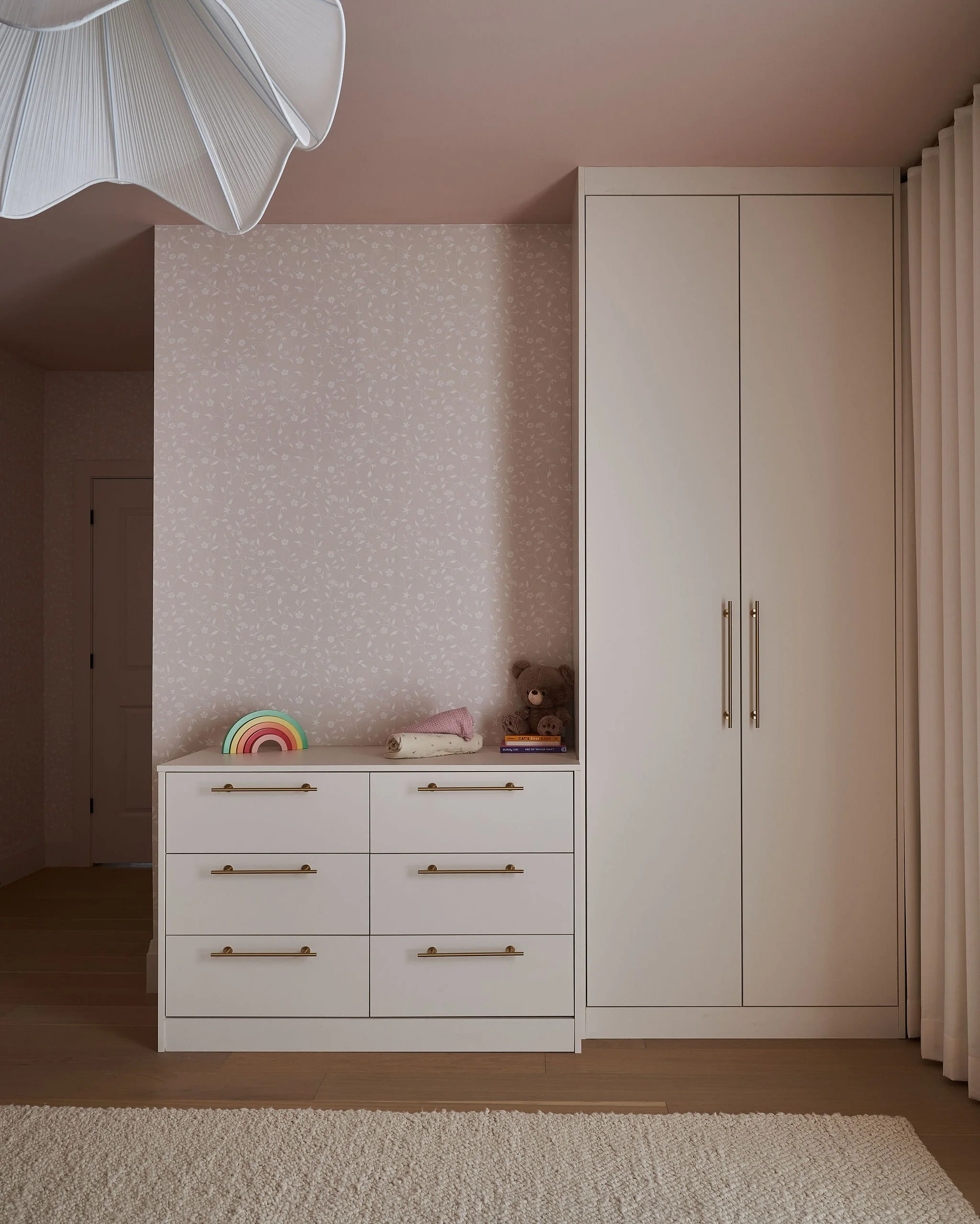 A white dresser with six drawers and gold handles next to a white wardrobe with two doors, against a pink wall with floral wallpaper. Items on the dresser include a rainbow decor, teddy bear, books, and a rolled towel. There is a beige plush rug on t