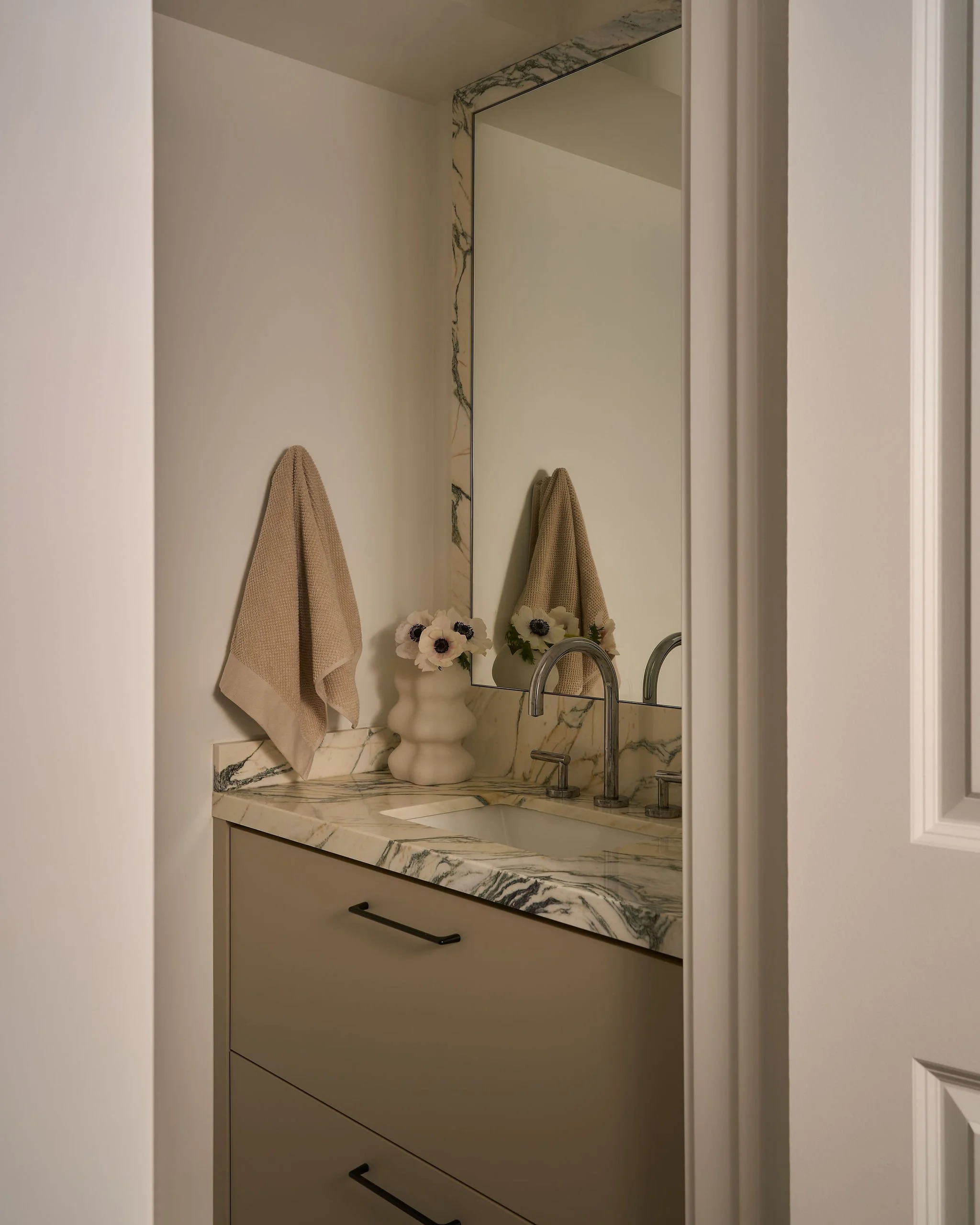 Bathroom vanity with marble countertop, a large mirror, beige hand towels hanging on each side, a white vase with white flowers, and a modern faucet.