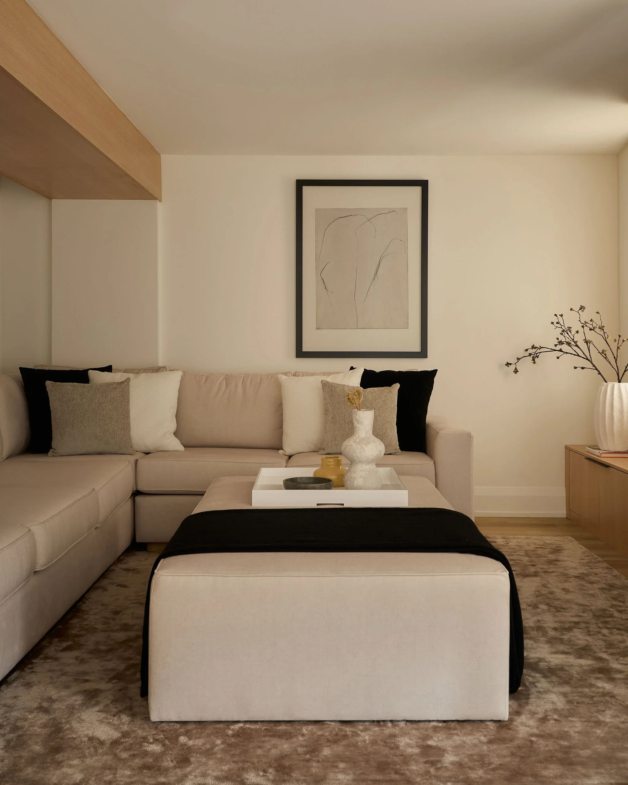 Living room with a sectional sofa adorned with beige, gray, and black pillows. A white coffee table in front holds decorative vases and bowls. Framed abstract line art hangs on the wall above. A plant with branches is on the right, and the room has a