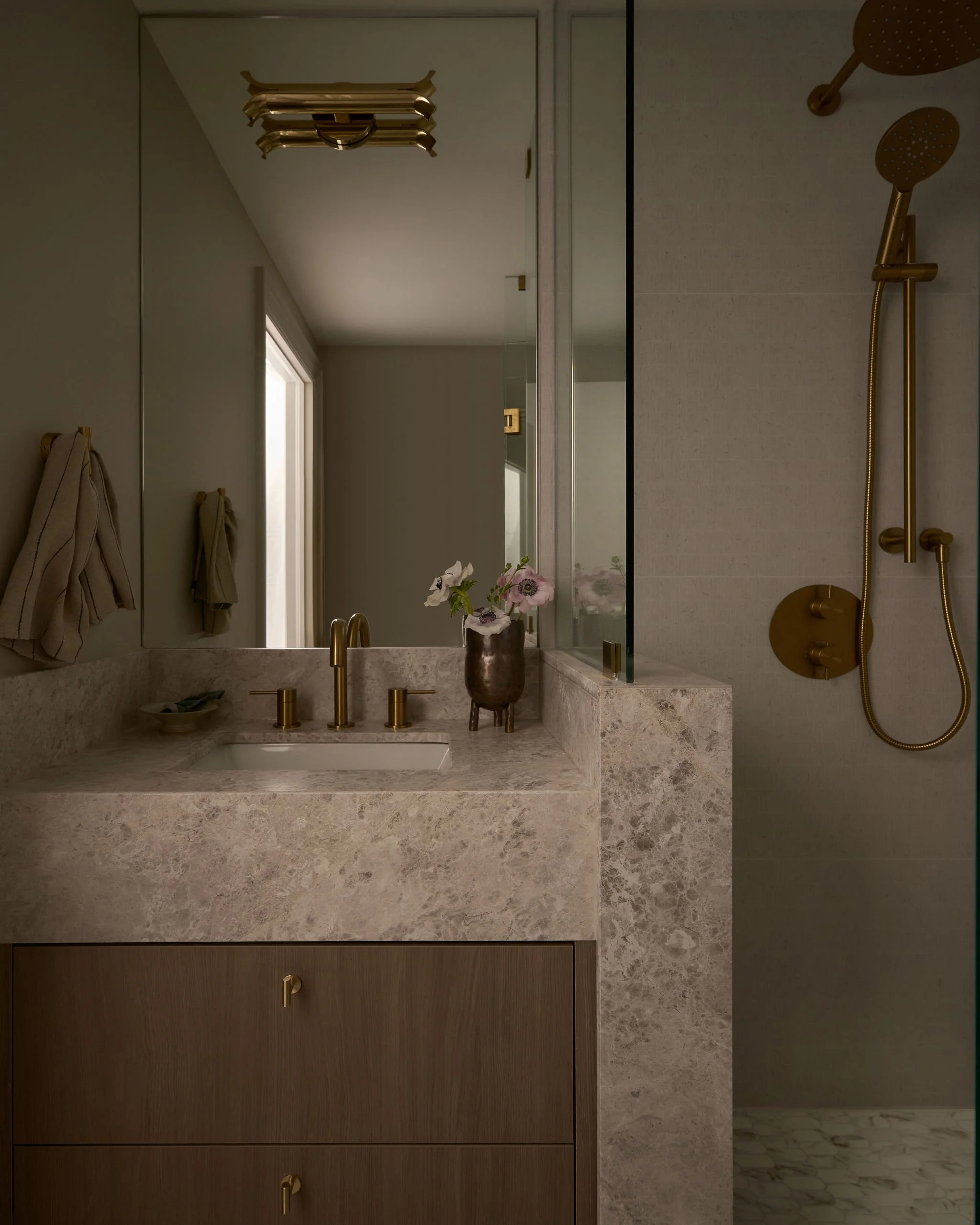 A bathroom with a marble vanity, gold fixtures, a black and gold vase with pink flowers, a mirror, beige towels on hooks, and a gold shower system with dual shower heads.