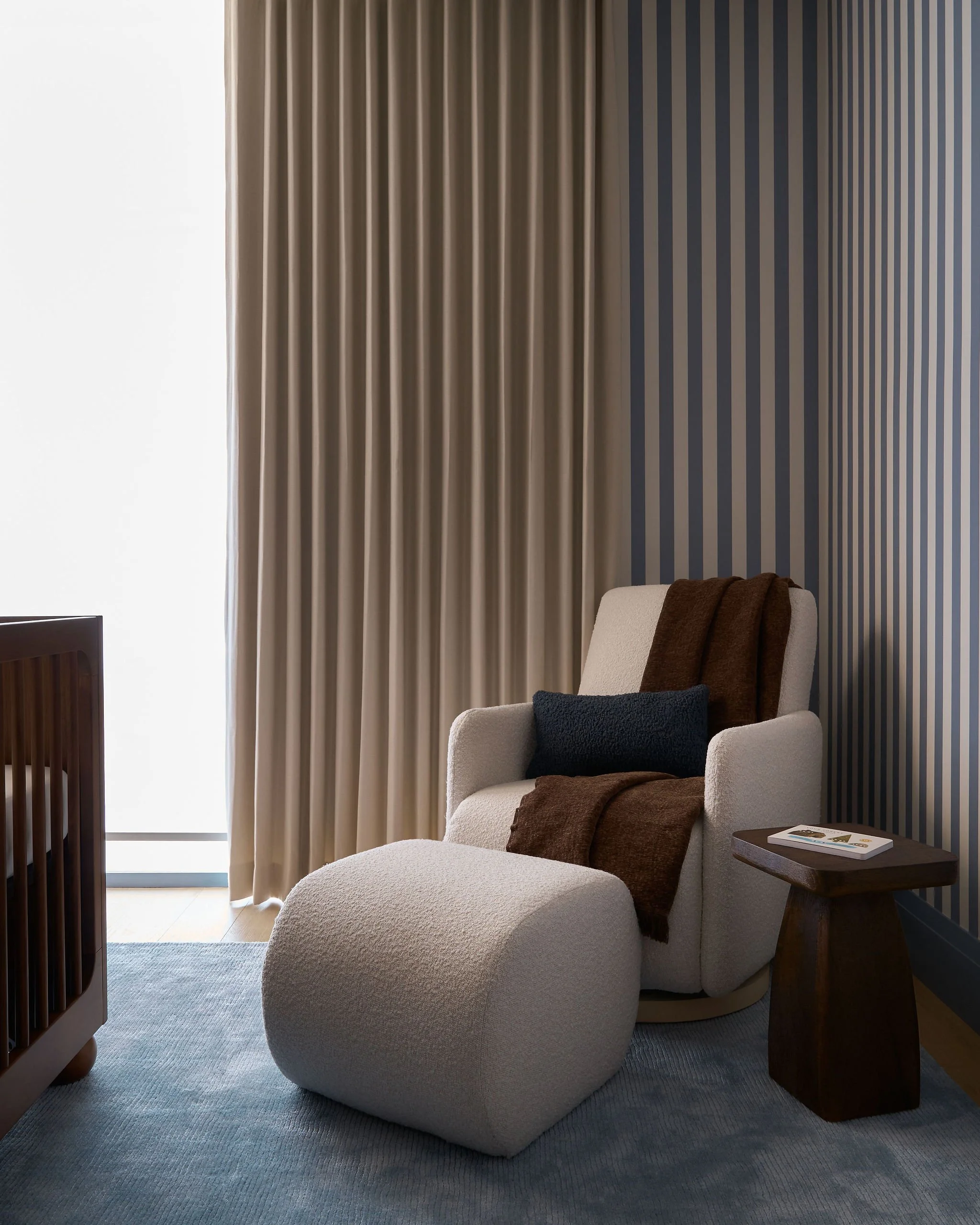 A cozy reading corner with a plush white armchair and matching ottoman, a dark wooden side table holding a magazine, and vertical beige and dark blue striped curtains letting in natural light.