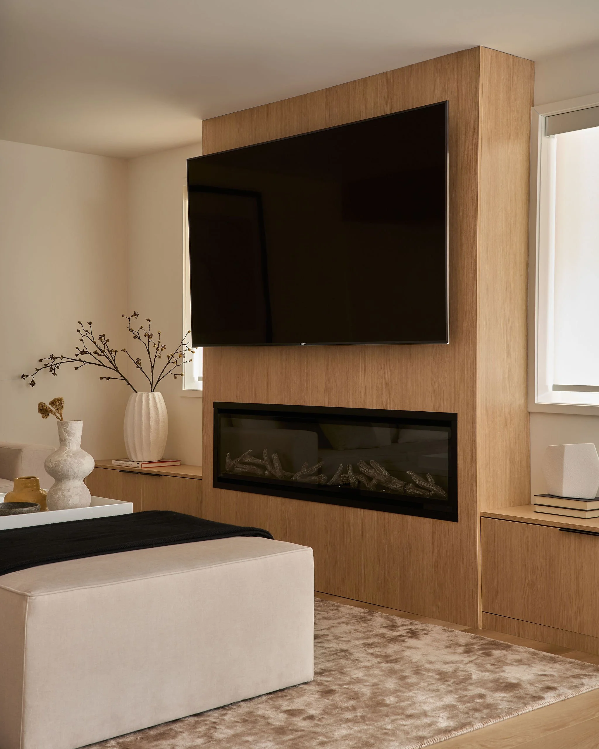 Living room with a mounted flat-screen TV on a wooden accent wall, decorated with a white vase containing dried branches, and a beige couch with a black throw blanket on a patterned area rug.