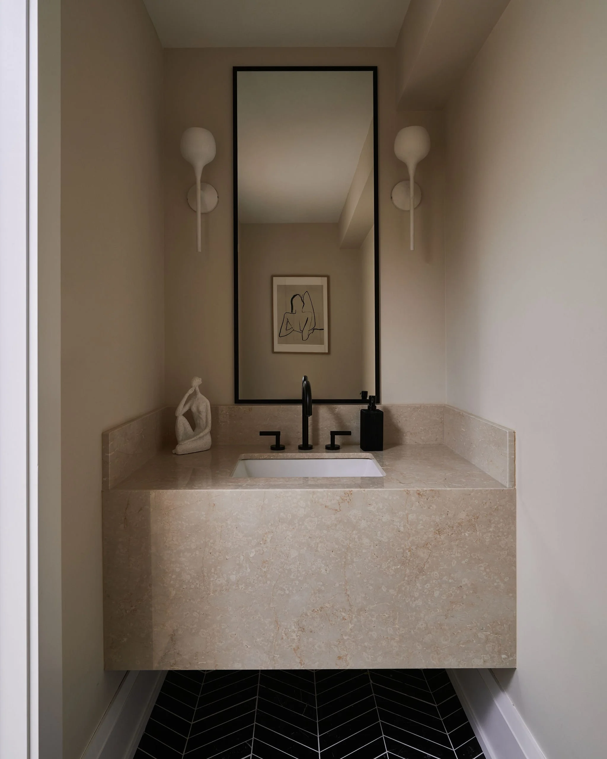 A bathroom vanity with a rectangular mirror, two white modern wall sconces on either side, a framed line drawing of a woman on the wall, a beige stone countertop with a built-in sink, a black faucet, and a small sculpture of a seated person on the le