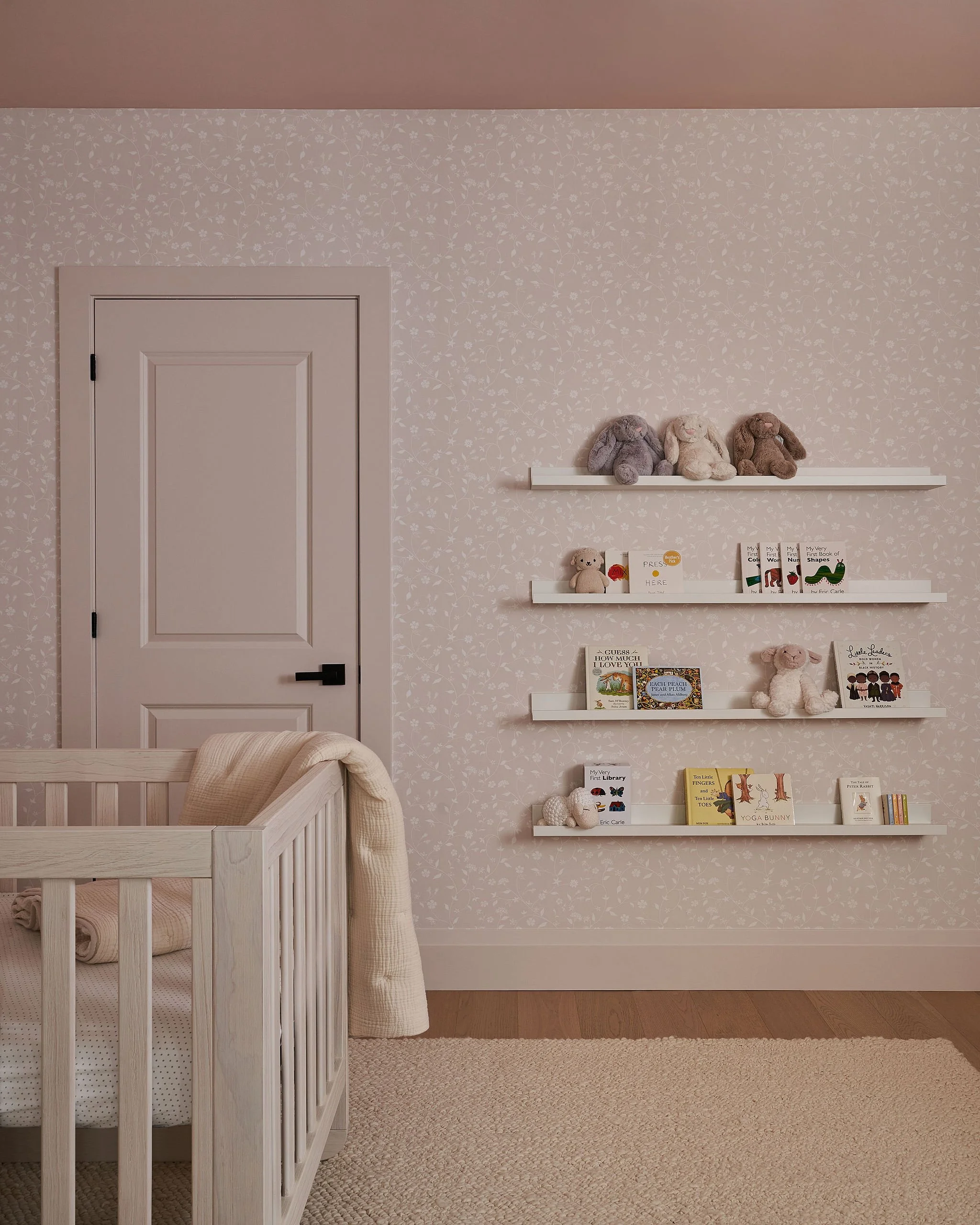 A children's nursery with a white crib, beige walls with floral wallpaper, and white shelves displaying stuffed animals and children's books.