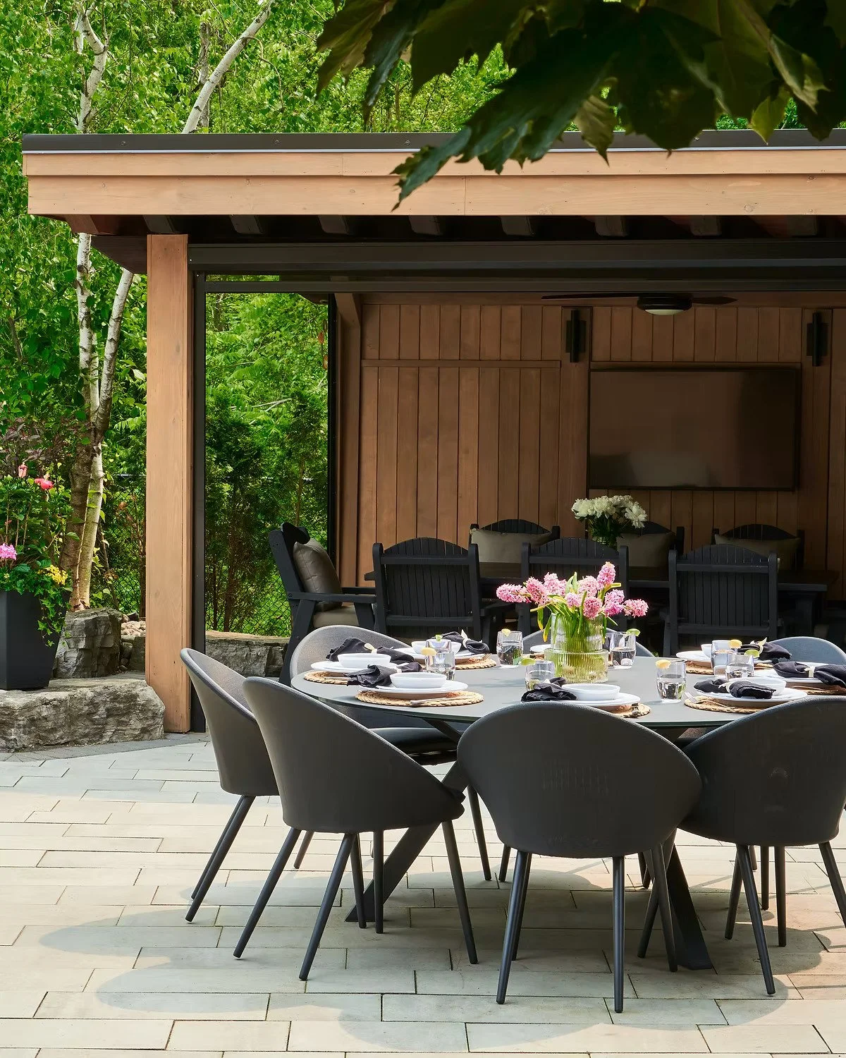 Outdoor dining area with a round table set for a meal, surrounded by black and grey chairs, and decorated with a centerpiece of pink flowers, with a wooded background and a wooden wall with a mounted television.
