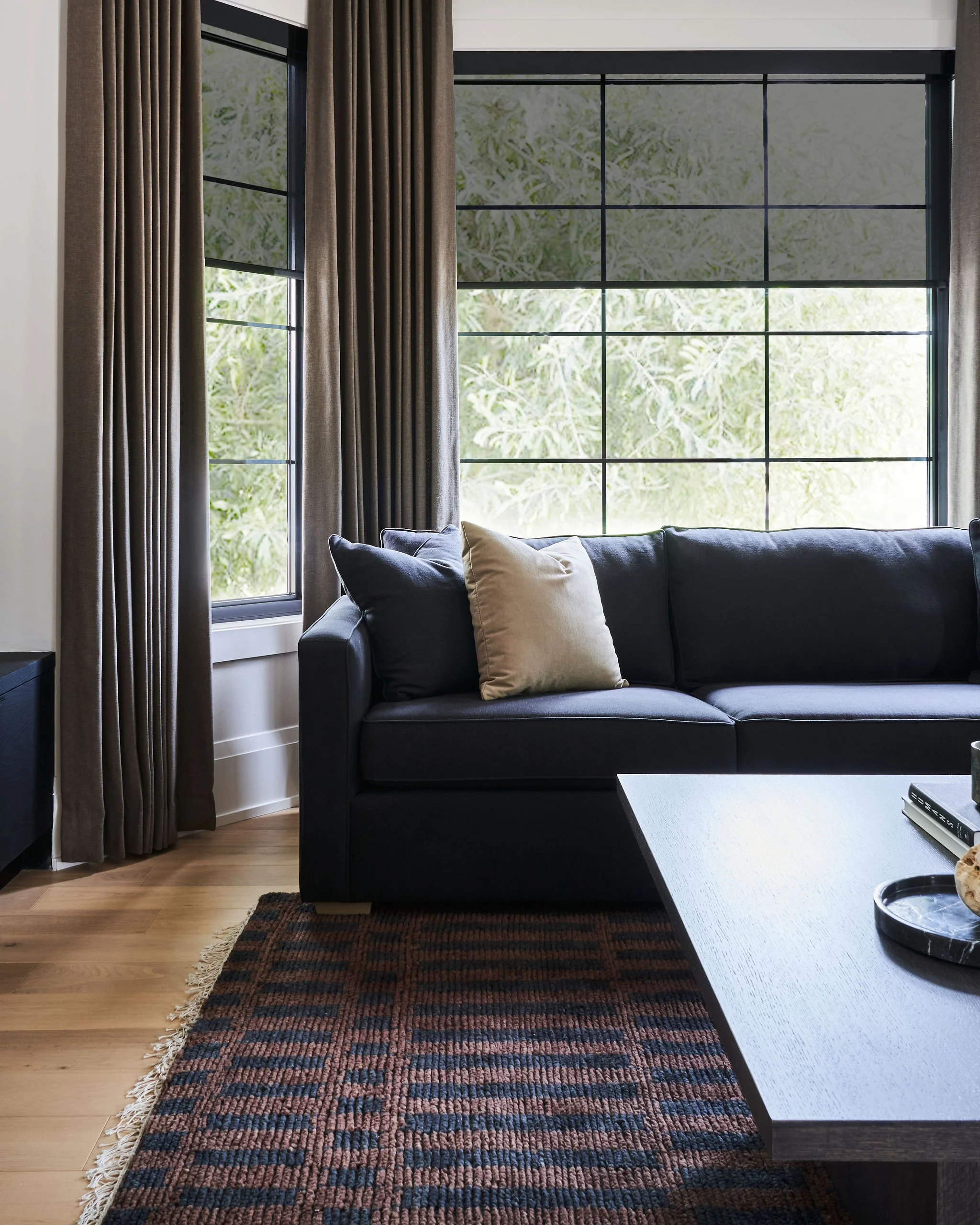 Living room with large grid-patterned window with black frames, beige curtains, a dark sofa with beige and black pillows, a light-colored wooden floor, a patterned rug, and a white coffee table with a book and a remote control.
