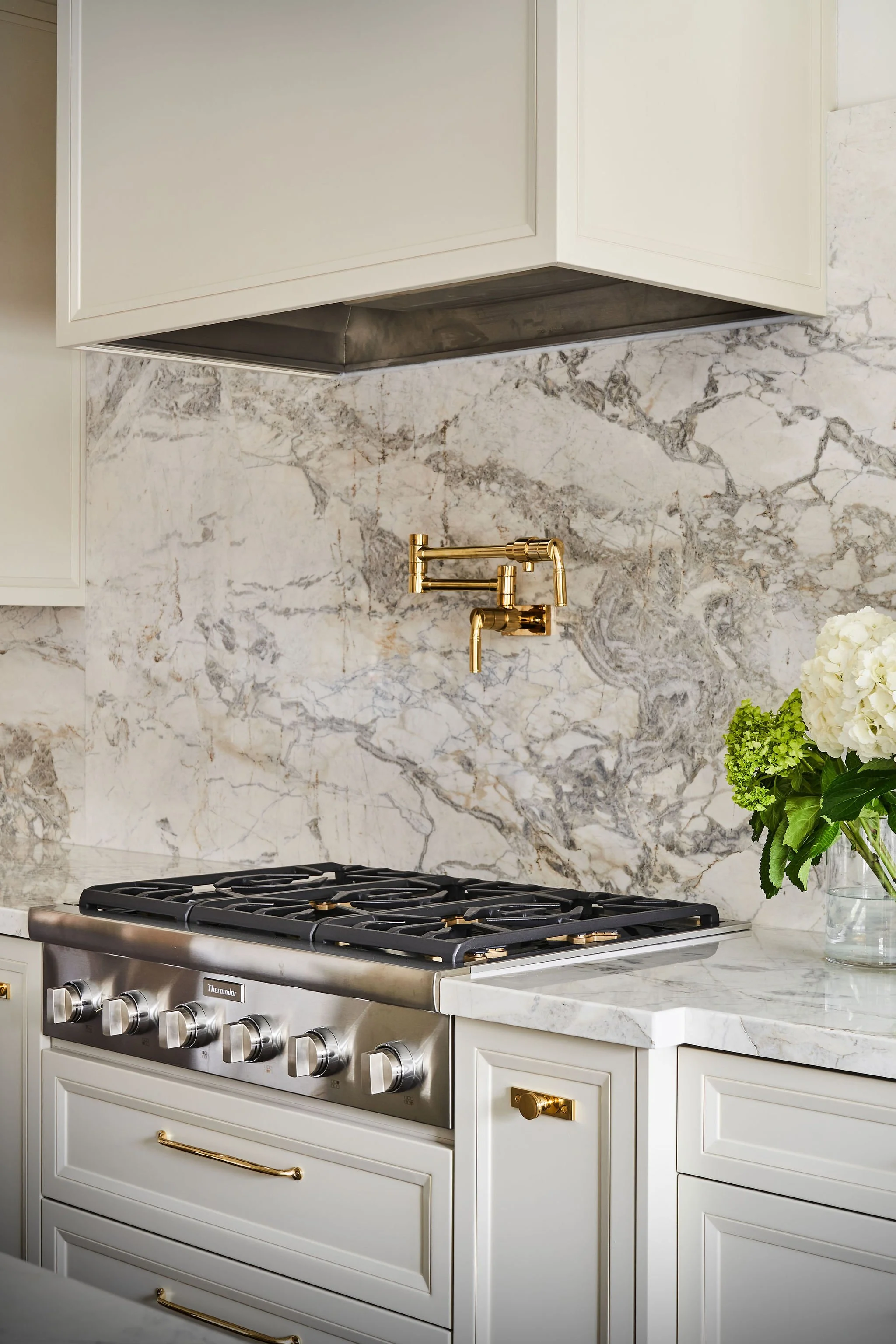 Close-up view of a modern kitchen with white cabinetry, a marble backsplash, a stainless steel gas stove, and a gold faucet with a bouquet of white flowers.