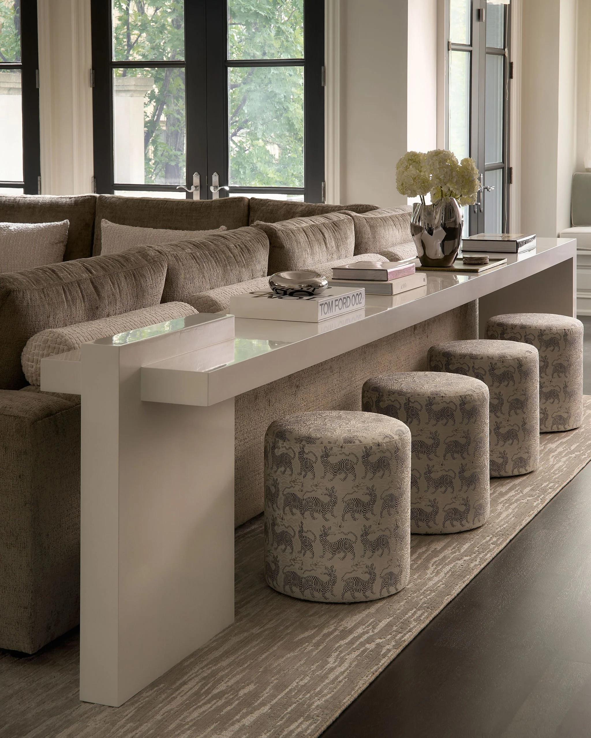 Living room with large windows, brown sectional sofa, white table with books, decorative bowl, flowers in a black vase, patterned ottomans, and a gray area rug.