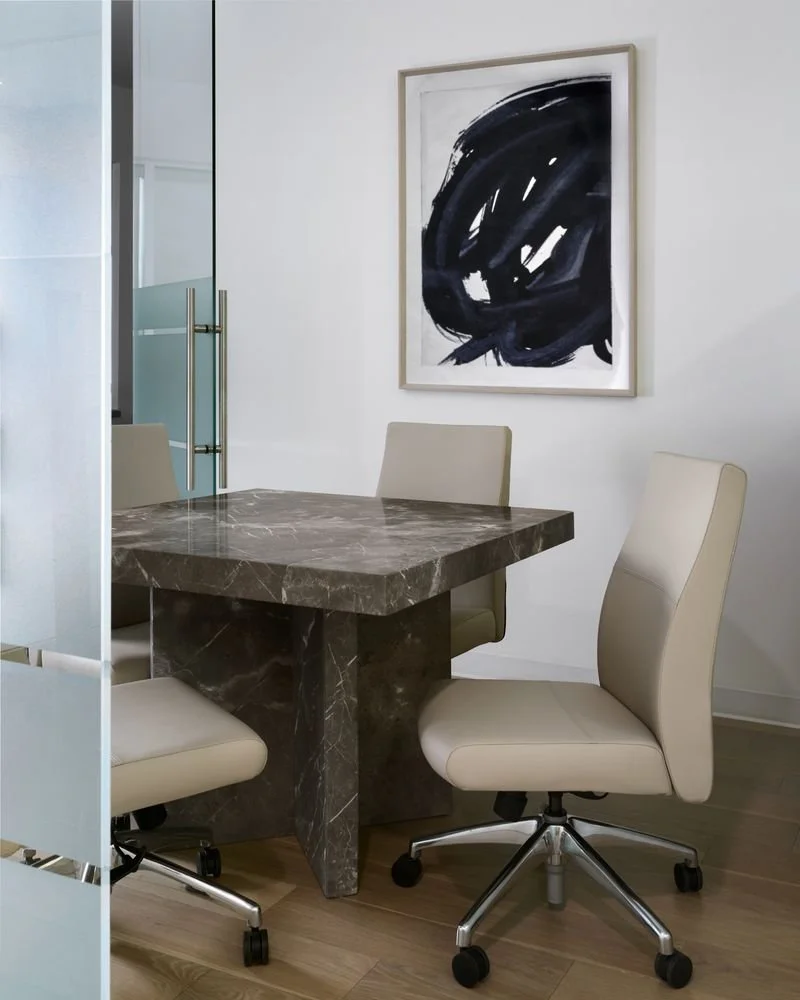 A modern meeting room with a marble table, three beige office chairs on casters, a white wall with an abstract black painting, and a sliding glass door.