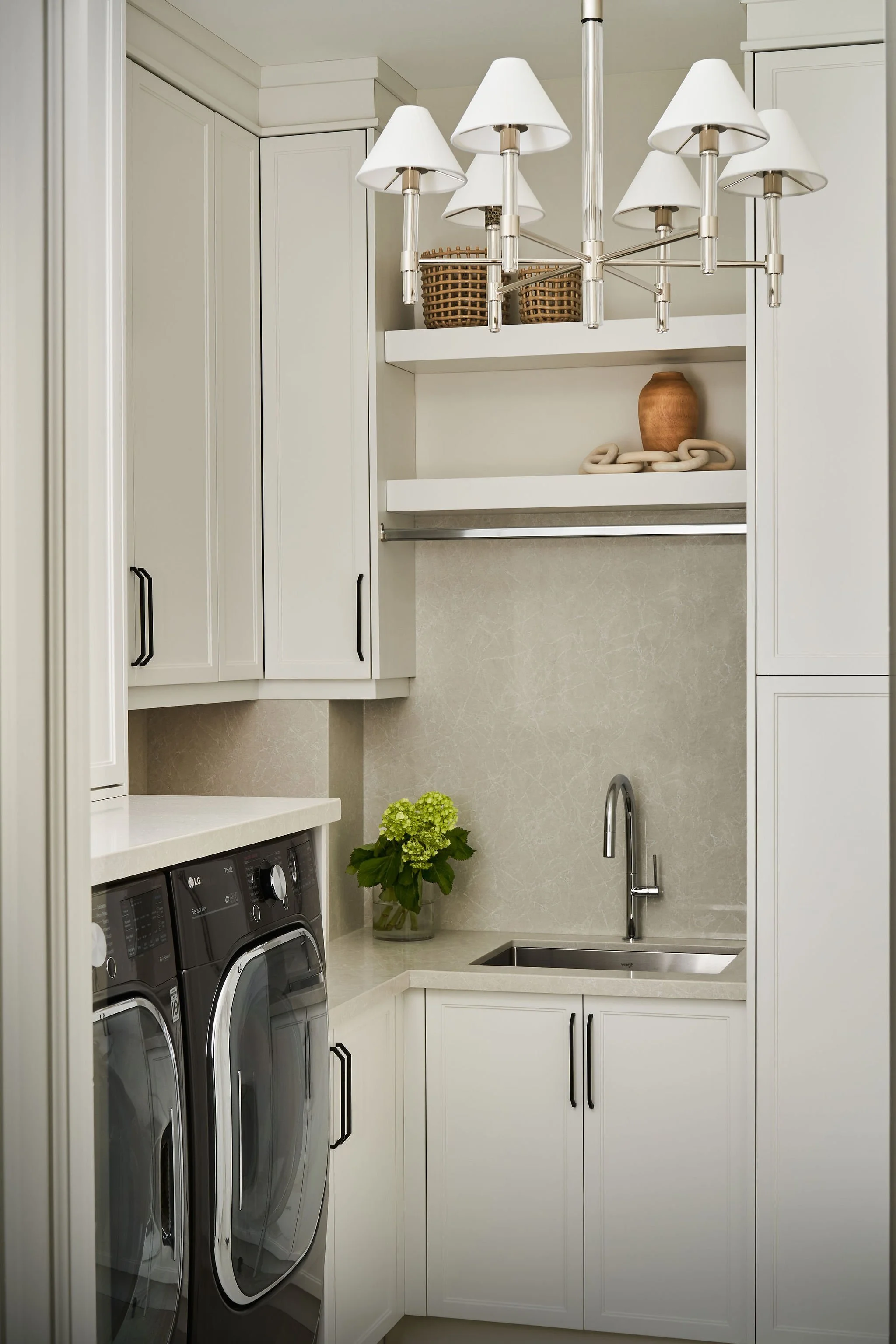 A laundry room with white cabinets, a small sink, and a countertop. There are two front-loading washing machines, a plant with green flowers, a modern chandelier, and a decorative vase with a chain on a floating shelf.