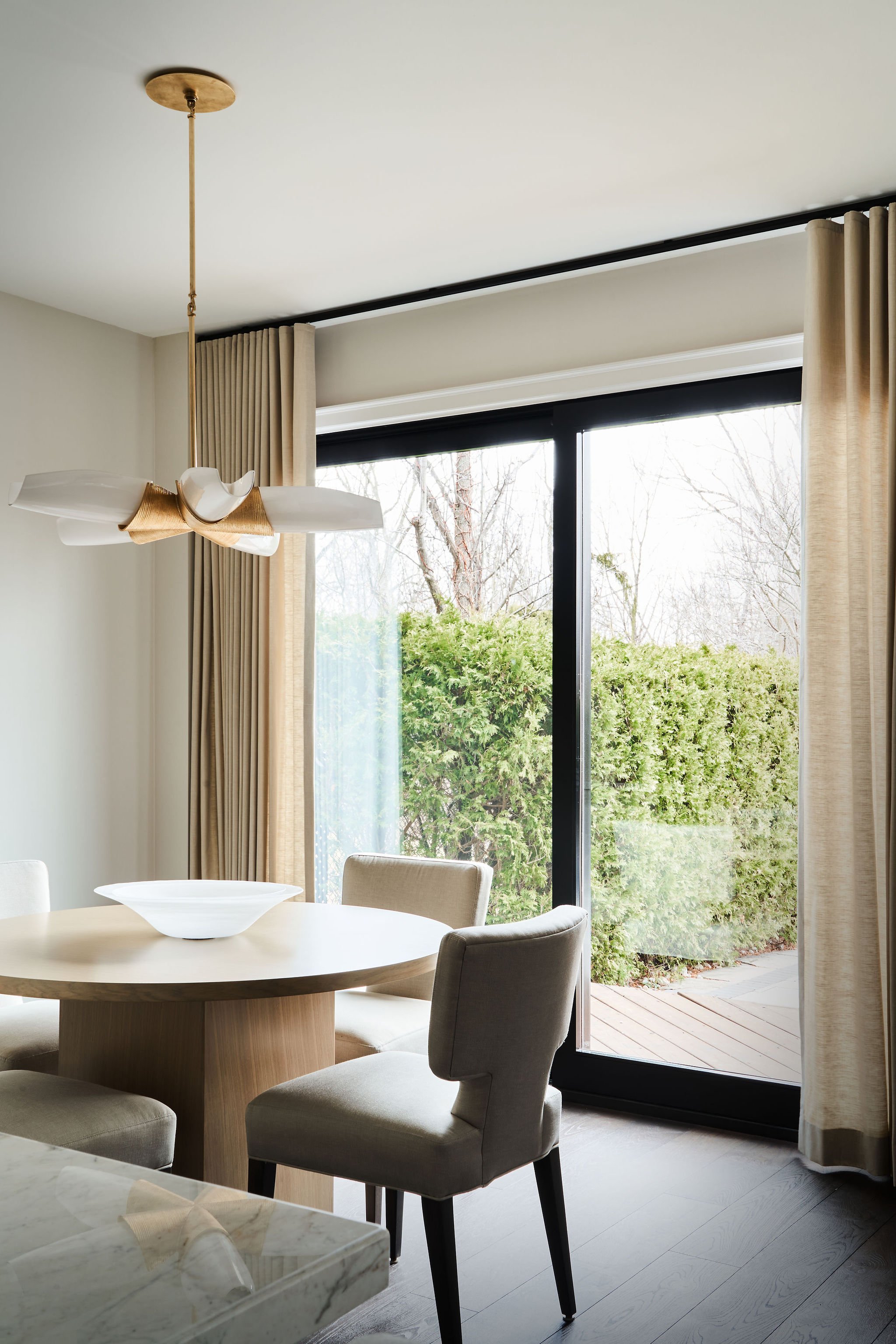 Modern dining room with large window, beige curtains, white chairs around a round wooden table, and a white decorative bowl. Outside, a patio and green bushes are visible.