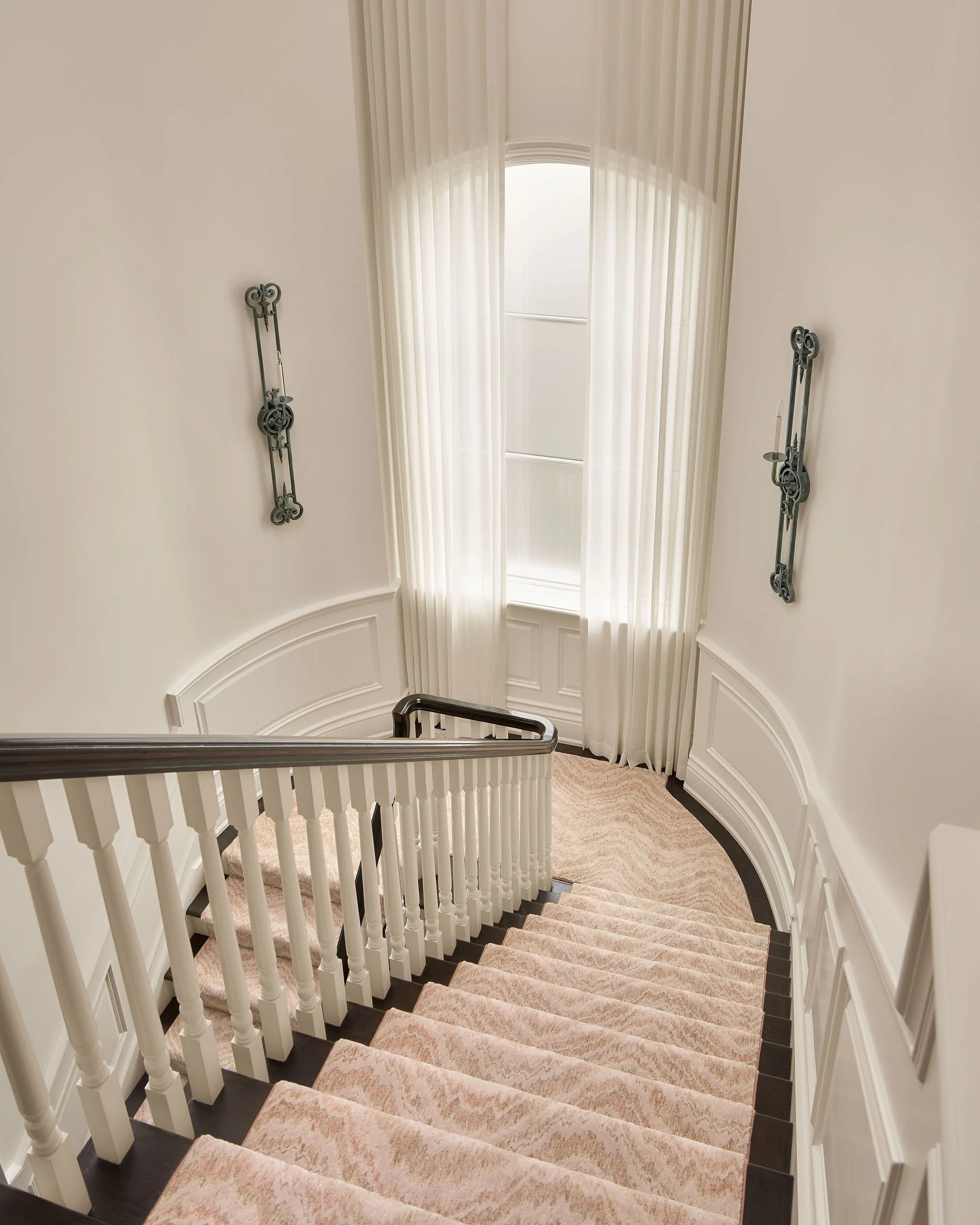 A staircase with beige carpeted steps, white railings, and a curved wall with paneled wainscoting. There are two decorative black wall hangings on the curved wall. An arched window with sheer white curtains is at the top of the stairs, allowing sunli