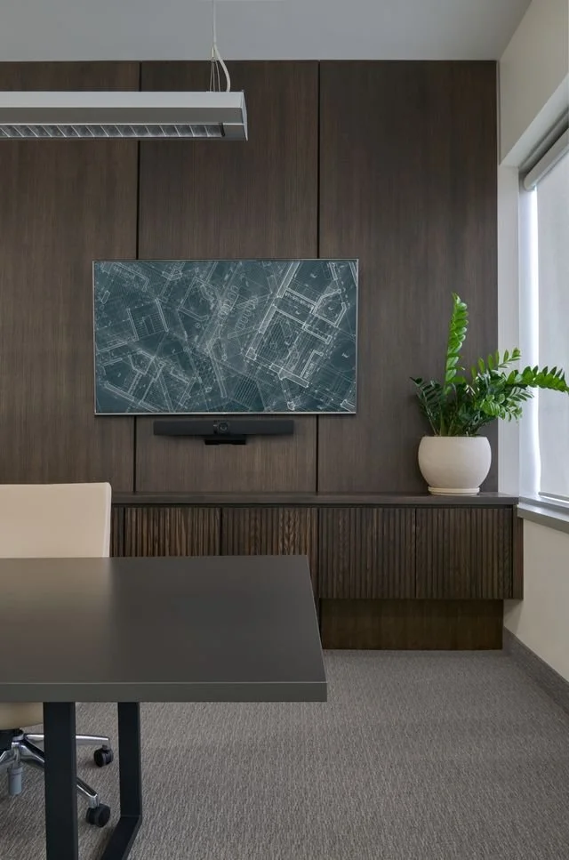 Modern conference room with a dark wood wall, a mounted screen displaying technical blueprint drawings, a white office chair, a black conference table, a potted plant, and a window with blinds.