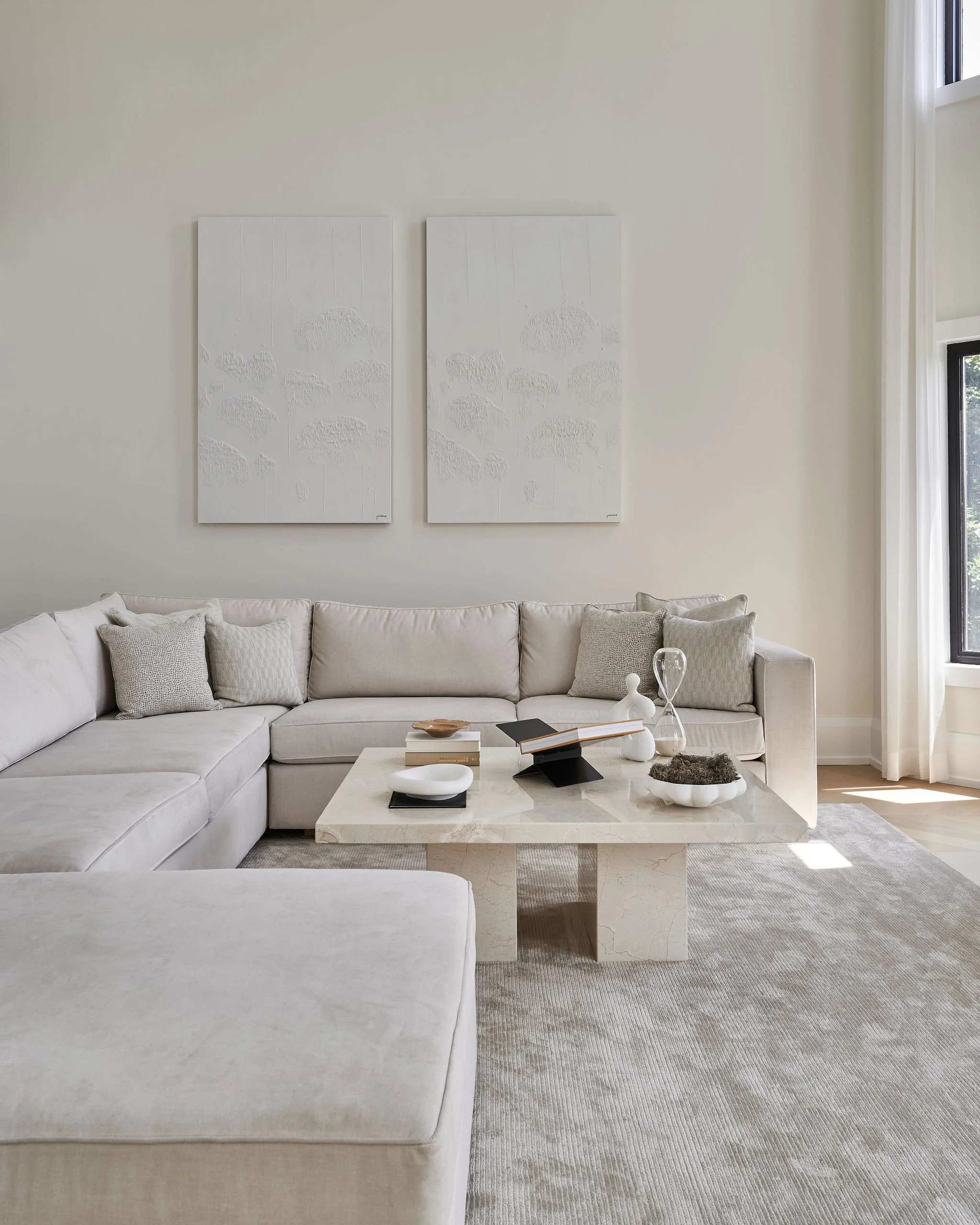 Minimalist living room with a white L-shaped sofa, pillows, a marble coffee table with decorative objects, wall art above, and natural light from a large window.