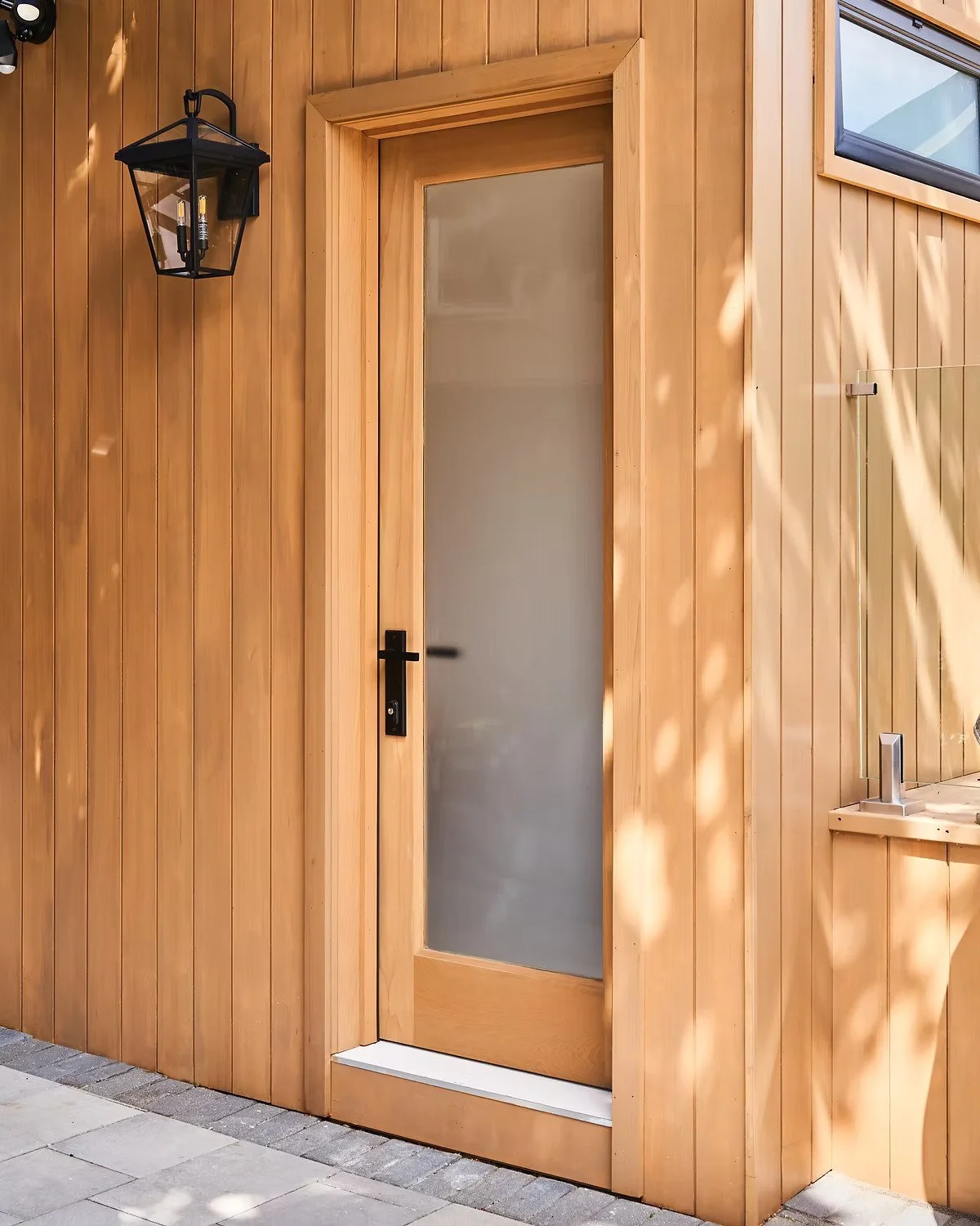 Wood-paneled exterior wall with a frosted glass door, black handle, black outdoor lantern light, and partial window.