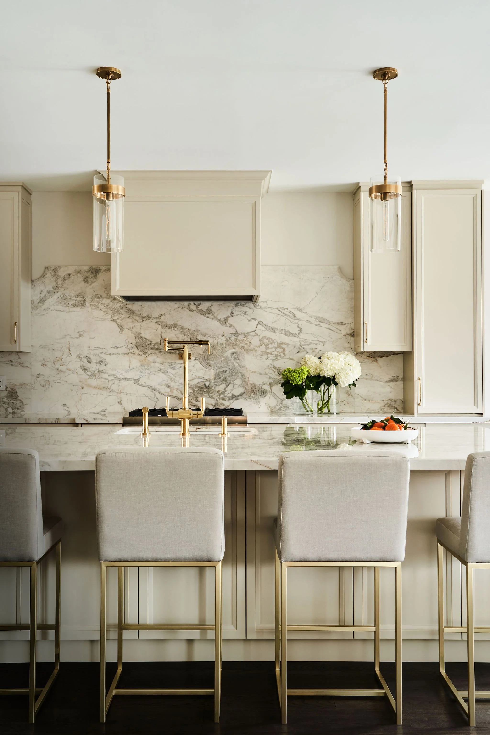 A kitchen with white cabinets, a marble backsplash, a white marble countertop, and gold accents, including a gold faucet and gold barstool legs. There are white chairs around a marble island, a bouquet of white flowers, and a bowl of oranges.
