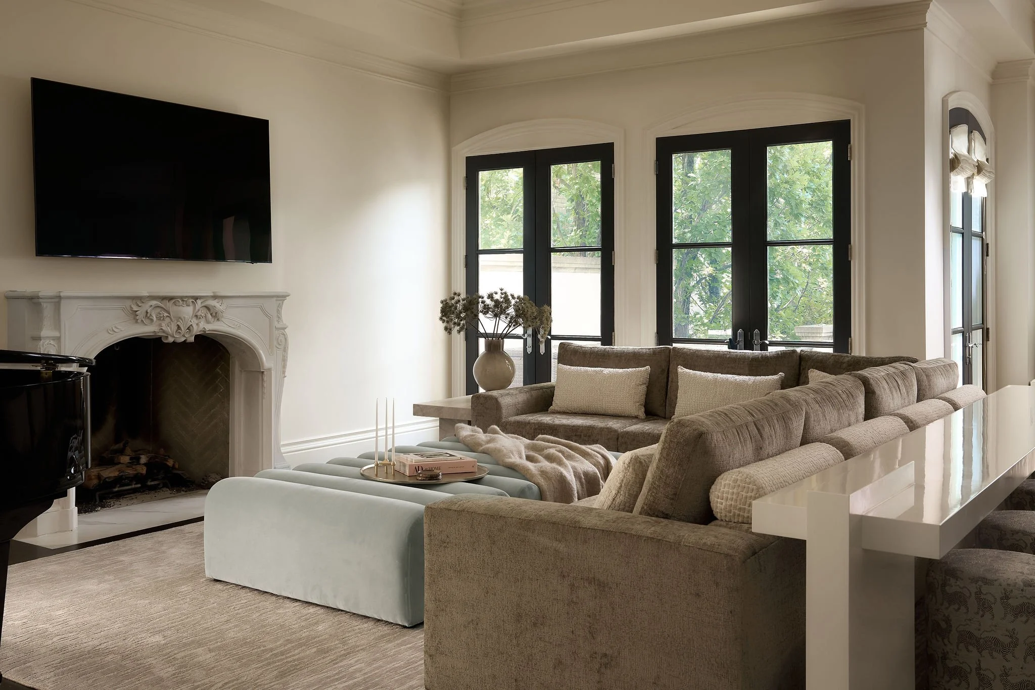 Living room with a large beige sectional sofa, a white marble coffee table with a decorative tray and candles, and a fireplace with ornate molding. There are large windows with black frames showing greenery outside and a mounted flat-screen TV above 