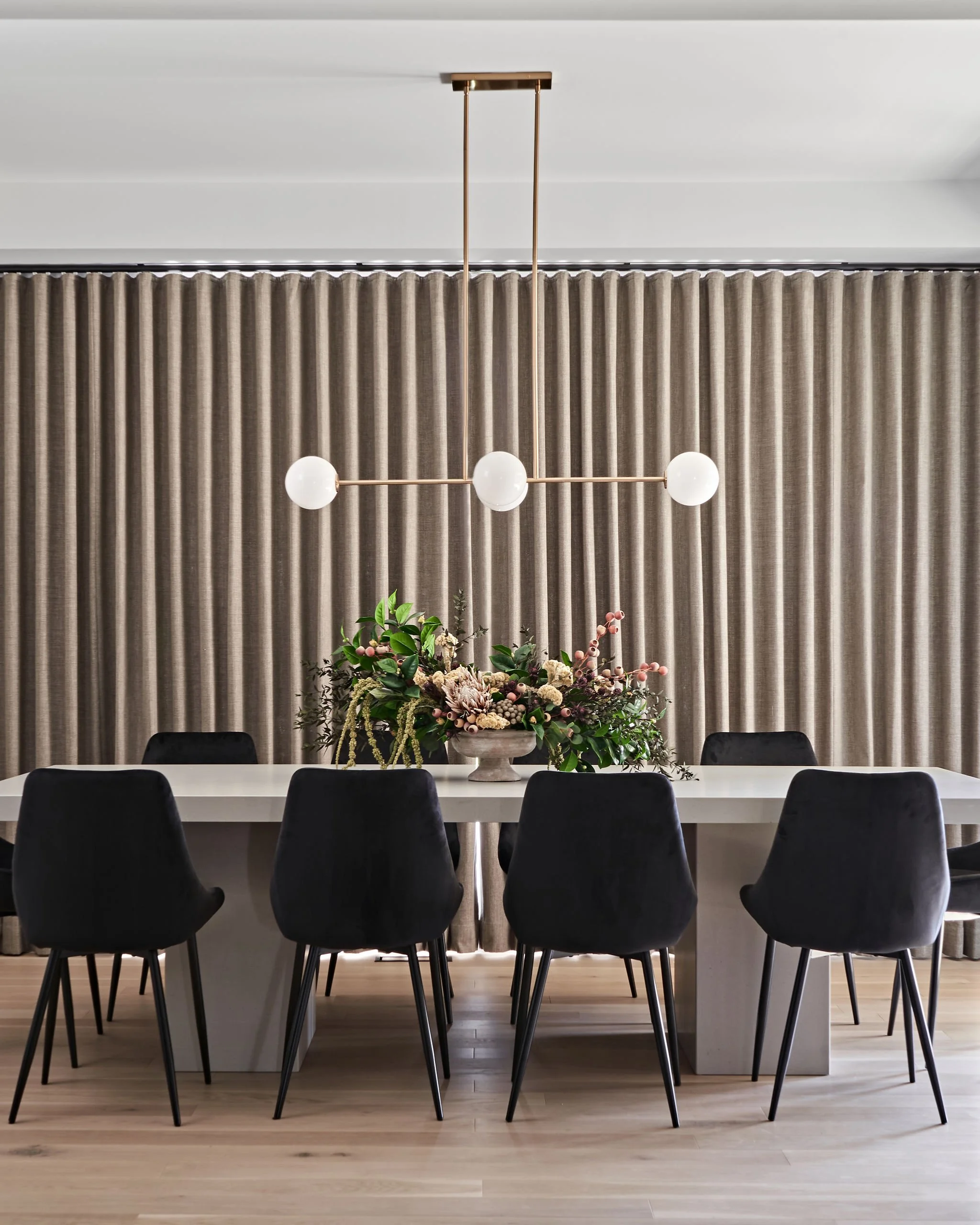 Modern dining room with a white table, six black chairs, a flower arrangement centerpiece, beige curtains, and a contemporary pendant light fixture with three white globes hanging from a gold rod.